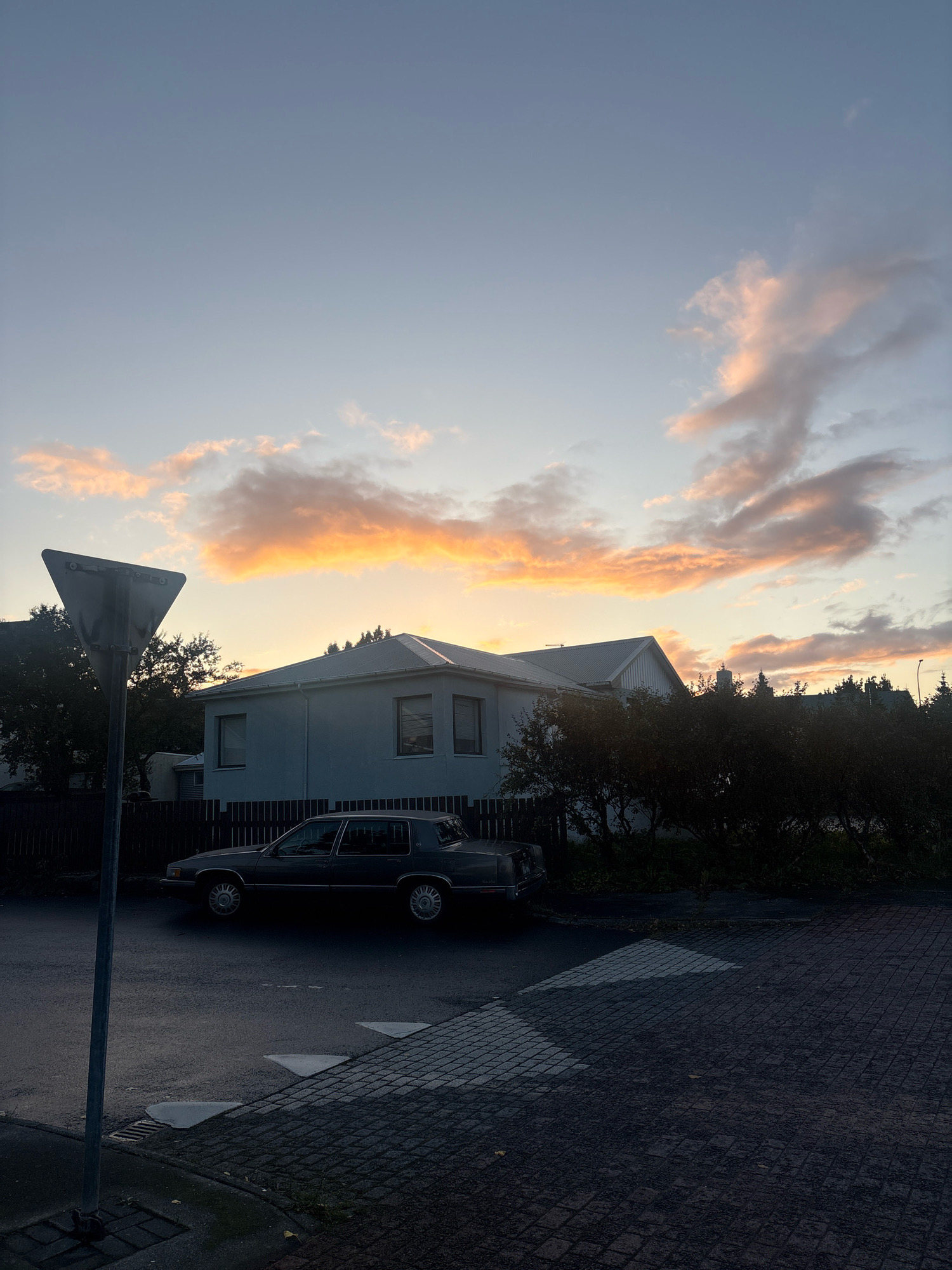 The image depicts a suburban street scene in Reykjavik, Iceland, during twilight. A light blue, single-story house with a white roof is visible on the right, partially obscured by dark foliage. A dark-colored sedan is parked on the street in front of the house. A yield sign on a metal pole stands in the foreground on the left. The sky is a gradient of pale blue to soft orange and pink, indicating the setting sun. The street is paved with asphalt, and a pedestrian crossing marked by white diagonal lines is present. Cobblestones line the edge of the sidewalk. The overall atmosphere is calm and quiet, characteristic of a residential area at dusk.