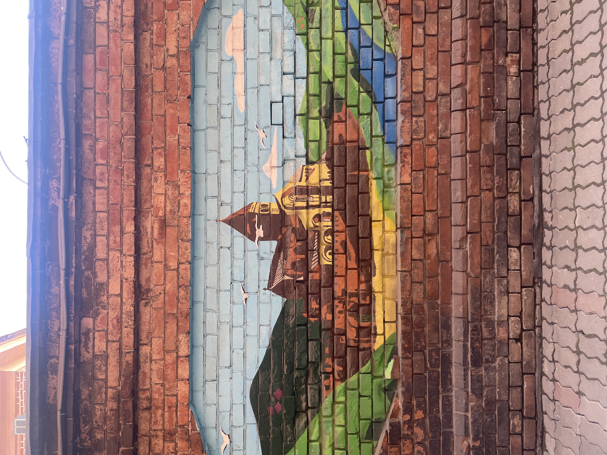 This is an image of a mural painted onto a brick wall. The mural depicts a scene with a building, possibly a church or cathedral, positioned within a landscape of trees and water. The sky is light blue with white streaks. There are two birds visible in the sky.
Based on the style of the building and the landscape depicted in the mural, it's likely the mural is depicting a scene from Krasnoyarsk, Russia.