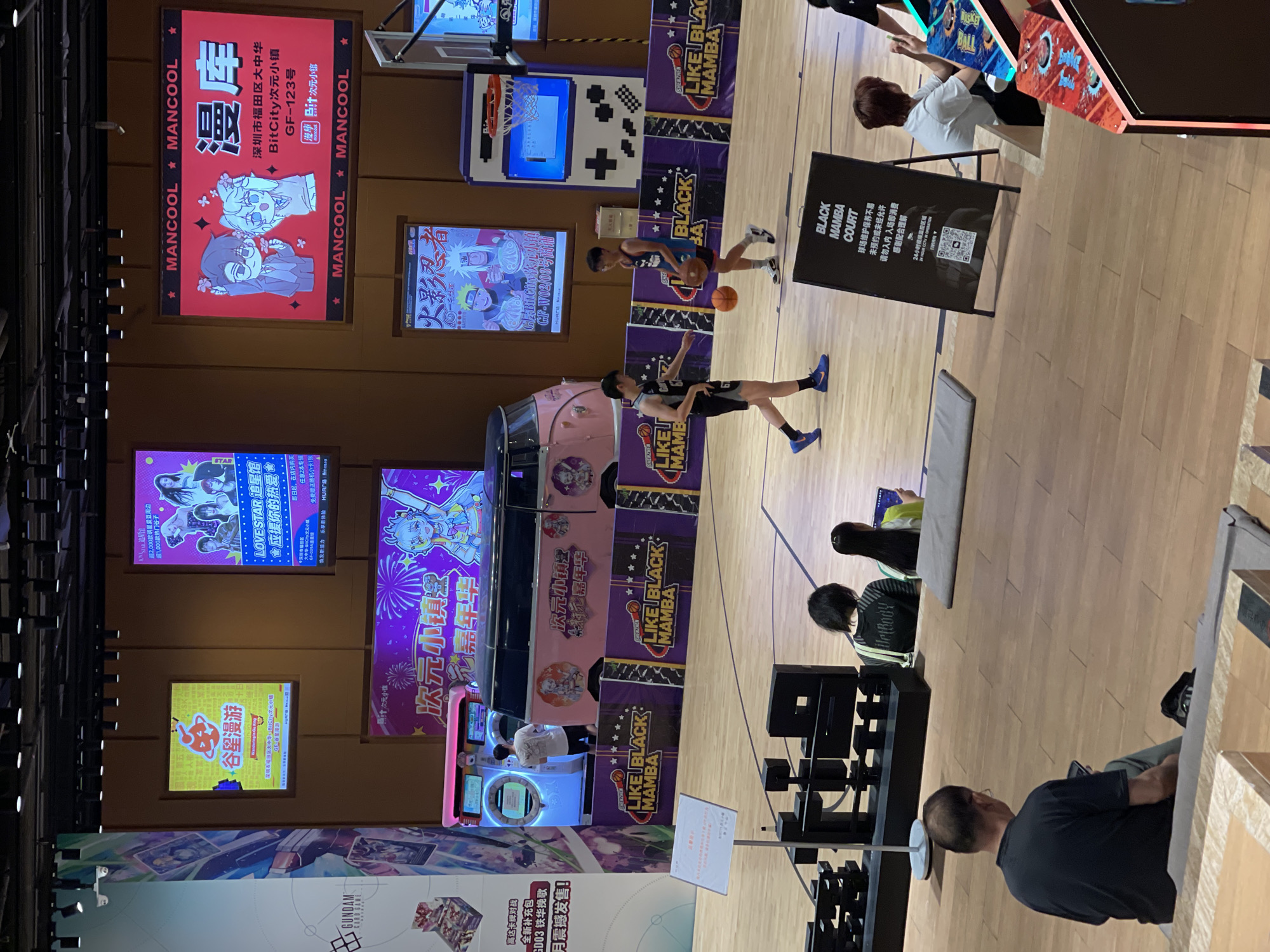 This media file depicts an indoor scene at what appears to be an entertainment or retail establishment, likely a game arcade or a themed store given the prominent displays and the presence of arcade machines. The setting is brightly lit, suggesting it's daytime or the interior is well-illuminated. The floor is made of polished wood, resembling a basketball court, and there are various colorful banners and screens advertising games and merchandise.
The central activity involves a few individuals engaged with the arcade games. Two young men are seen playing a basketball-themed game on a court-like area. Other people are visible, some watching the game, others interacting with the machines, and one person appears to be seated, possibly observing or resting. The overall atmosphere seems casual and engaging.
Several pieces of text are visible throughout the image, primarily in Chinese. These include advertisements for "MANCOOL," "LOVE STAR 追星馆," "次元小镇," and "GUNDAM CARD GAME." There are also signs with game titles like "火影忍者" (Naruto) and mentions of "BLACK MAMBA COURT" with instructions for entry. The text provides clues about the location being in Futian District, China, specifically at BitCity, a "次元小镇" (dimensional town or anime town). The presence of anime and game-related imagery suggests a focus on pop culture and entertainment.