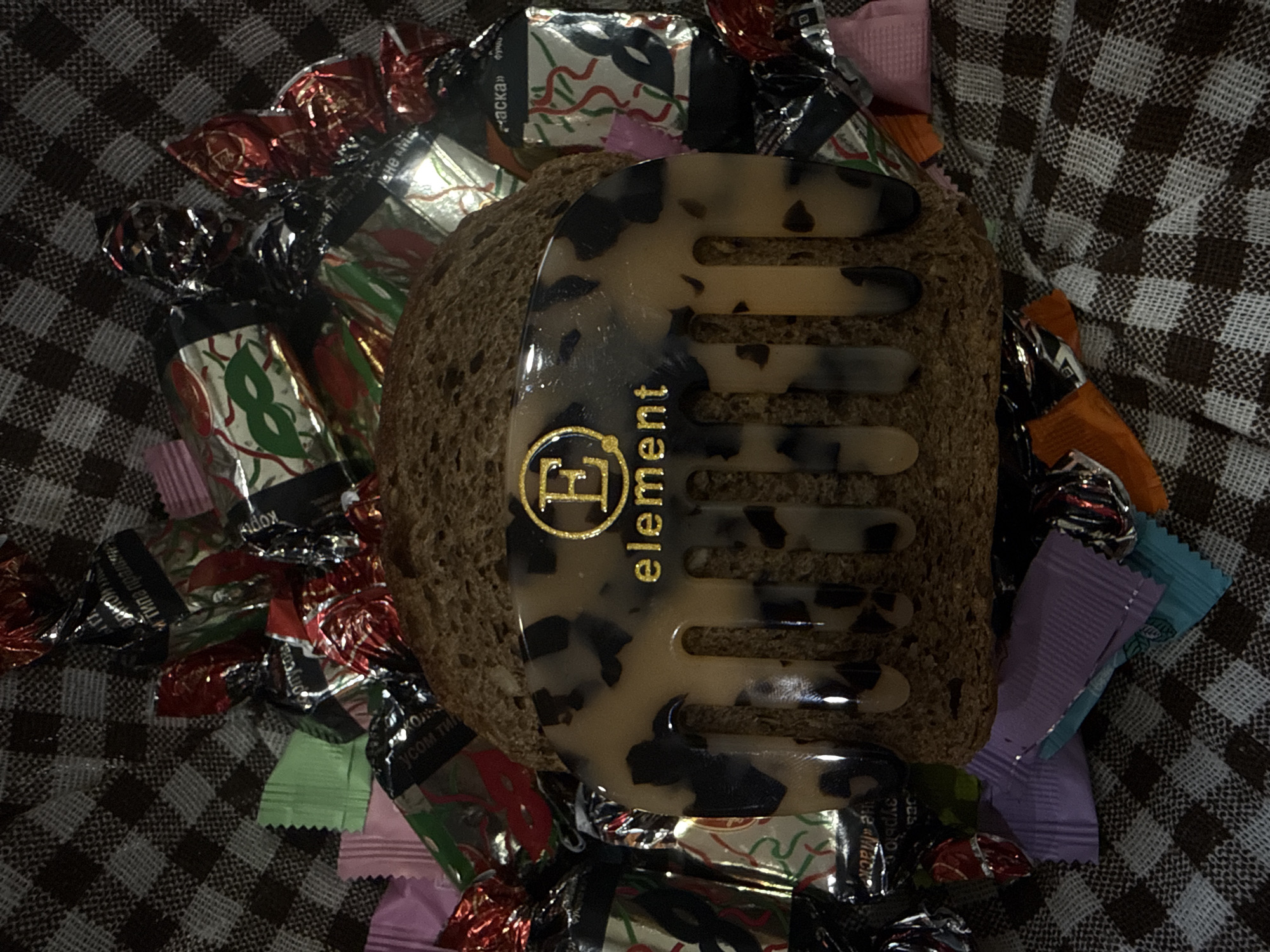 The image presents a still life composition on a brown and white checkered surface, possibly a tablecloth or blanket. A slice of dark bread serves as the base for a hair comb, beige with dark spots, and a gold-colored "Element" logo printed on it. The scene is surrounded by an assortment of wrapped candies, adding color and variety to the setting. No people are visible in the frame. Location is not verifiable from the image.