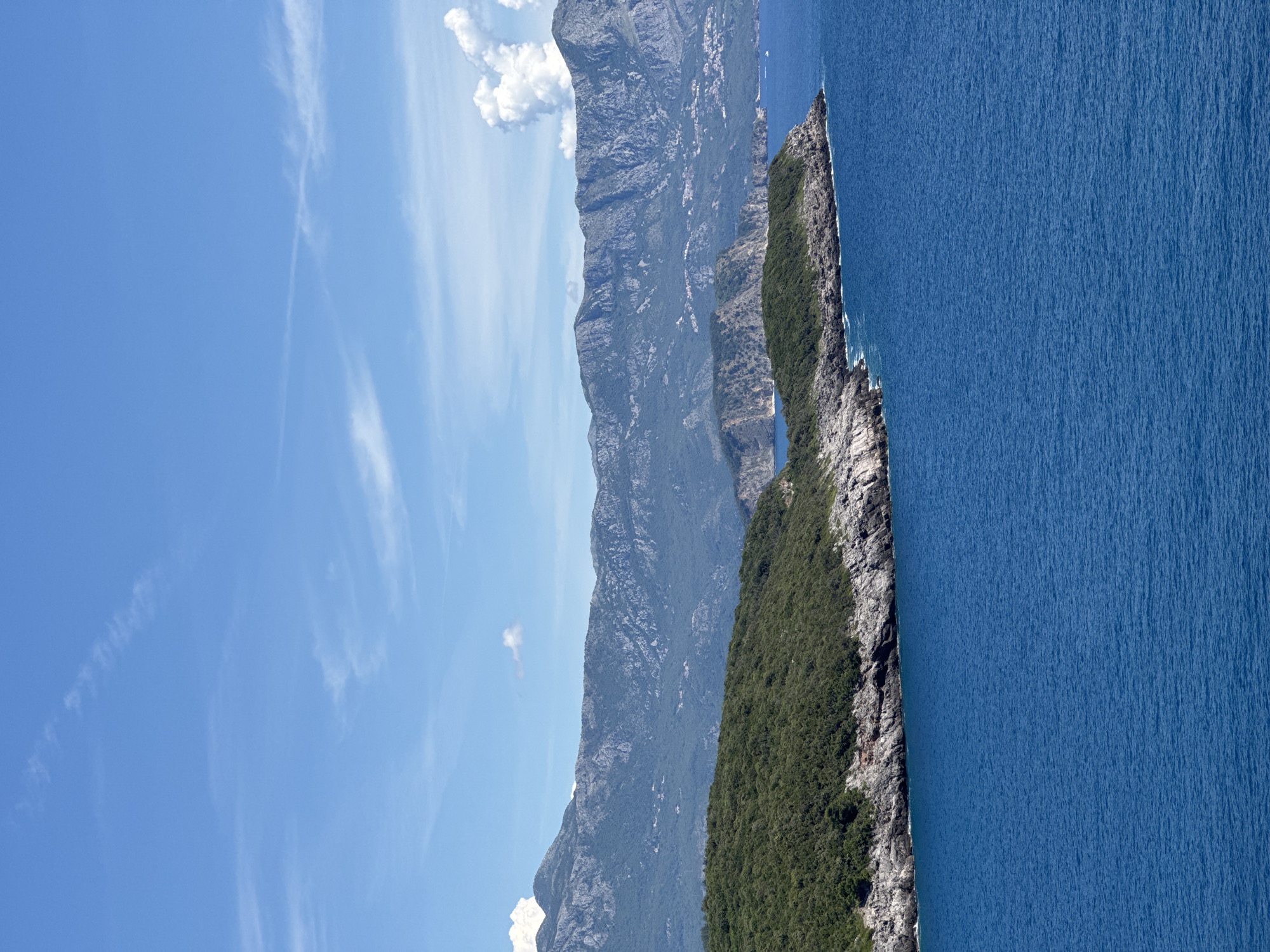 This image captures a serene coastal landscape in Dubraljevina, Montenegro. The foreground is dominated by the deep blue Adriatic Sea, its surface rippled by a gentle breeze. A rocky, tree-covered peninsula juts out into the sea from the left side of the frame. Beyond the peninsula, the coastline continues with more rocky outcrops and lush vegetation, leading to the distant, majestic mountains that form the backdrop of the scene. The mountains are rugged and appear to be covered in a mix of rocky terrain and dark green vegetation. Wispy white clouds drift across a clear blue sky, casting subtle shadows on the landscape below. No people or specific man-made structures are prominently visible in this image, emphasizing the natural beauty of the location.