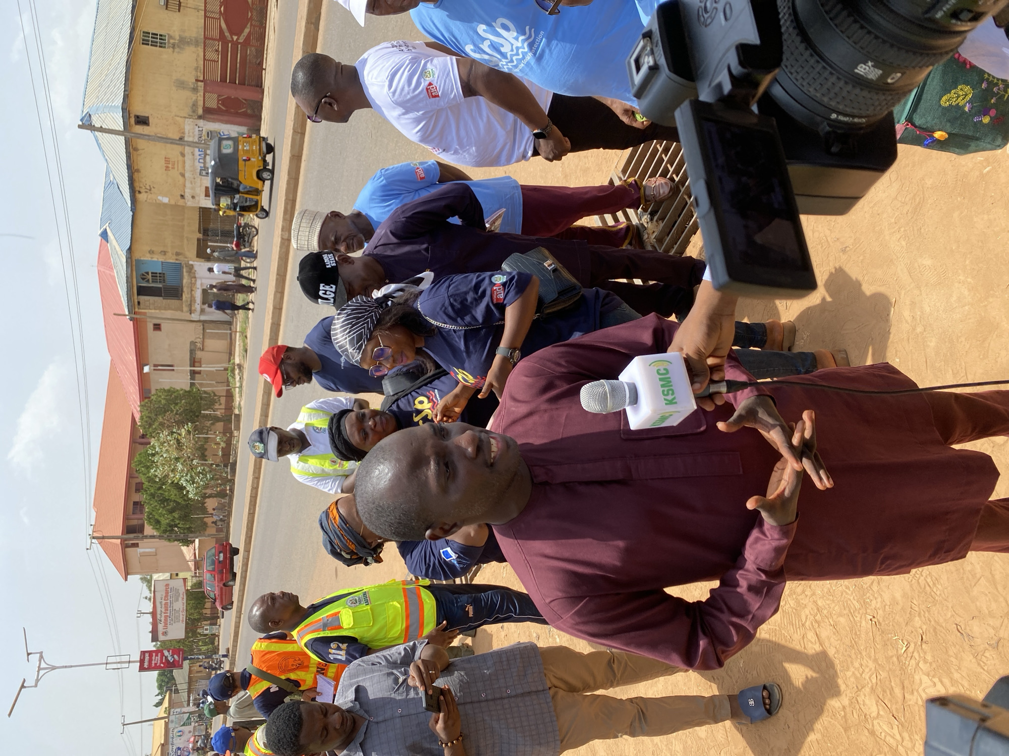 A man in a burgundy shirt is being interviewed by a reporter holding a microphone labeled "KSMC". The interview takes place on a street in Sabon Tasha, Nigeria, surrounded by a crowd. Background details include a yellow auto-rickshaw, and the "Living Faith Church" is identifiable, along with signs related to environmental and social protection. The overall scene suggests a public interest story or event.