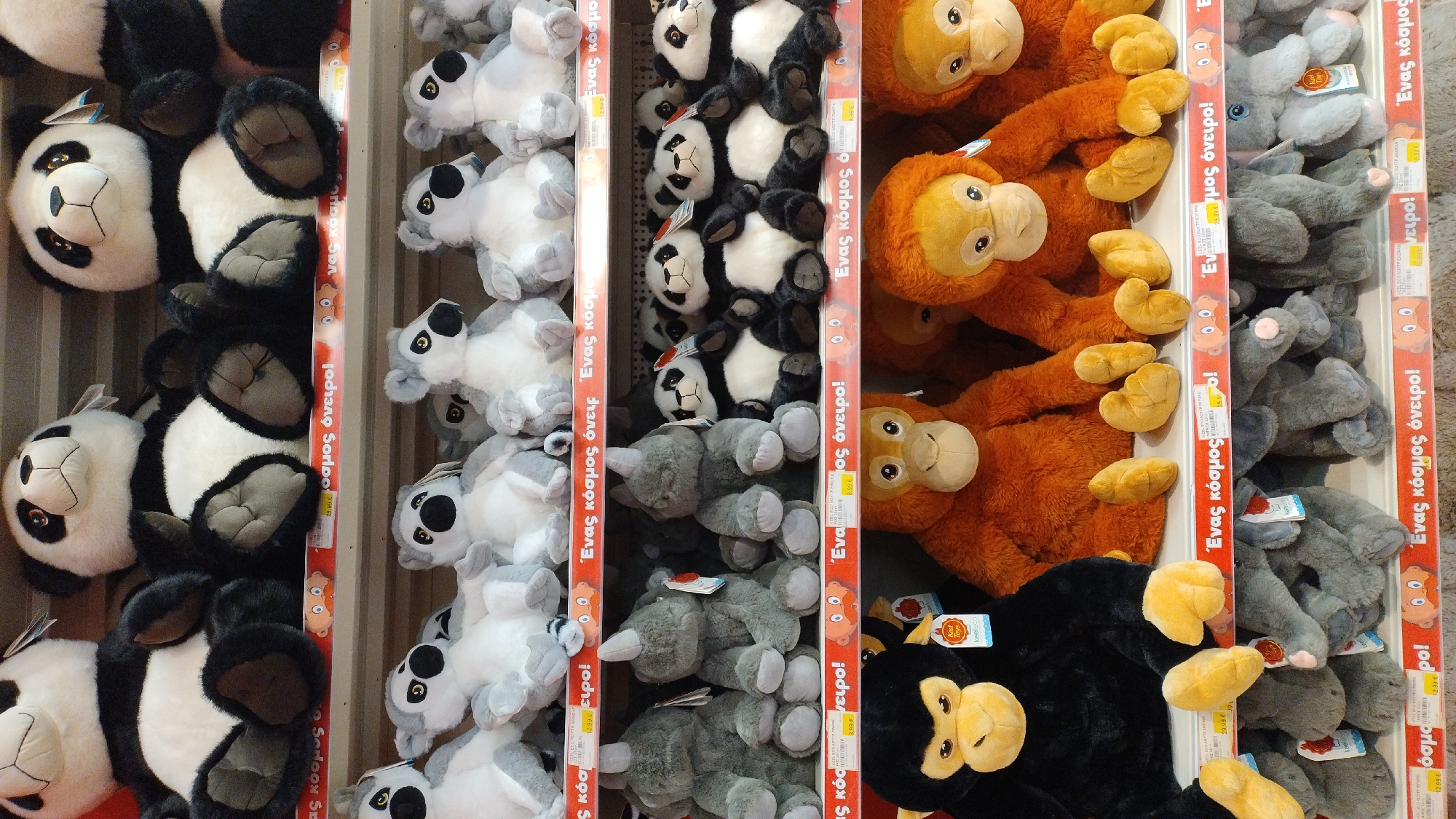 The image shows a retail display of stuffed animals in a store. The stuffed animals are arranged on shelves and include pandas, monkeys, orangutans, and elephants. The store appears to be a toy store or a department store with a toy section. The lighting is bright and evenly distributed, suggesting an indoor setting. The shelves are made of metal and are filled with plush toys. There are price tags visible on some of the toys, which are in Euros, suggesting the location is within the Eurozone. The writing on the price tags and promotional materials appears to be in Greek. The specific location is the Jumbo store in Pylaia Municipal Unit, Greece, as indicated by the Greek text which translates to "Jumbo is a world of dreams".