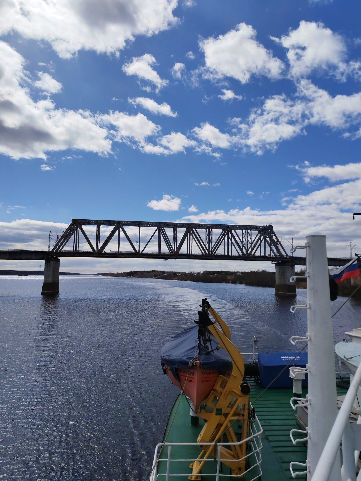 A medium shot shows a steel truss bridge crossing a wide body of water, as seen from the deck of a ship. The bridge has a dark, rust-colored appearance. The sky is a bright blue with scattered white clouds. The water has a dark blue hue with slight ripples and reflections. The ship's deck is green and contains a yellow life raft davit, a blue container, and a white railing with a Russian flag attached. The location is a river. The city is unknown.