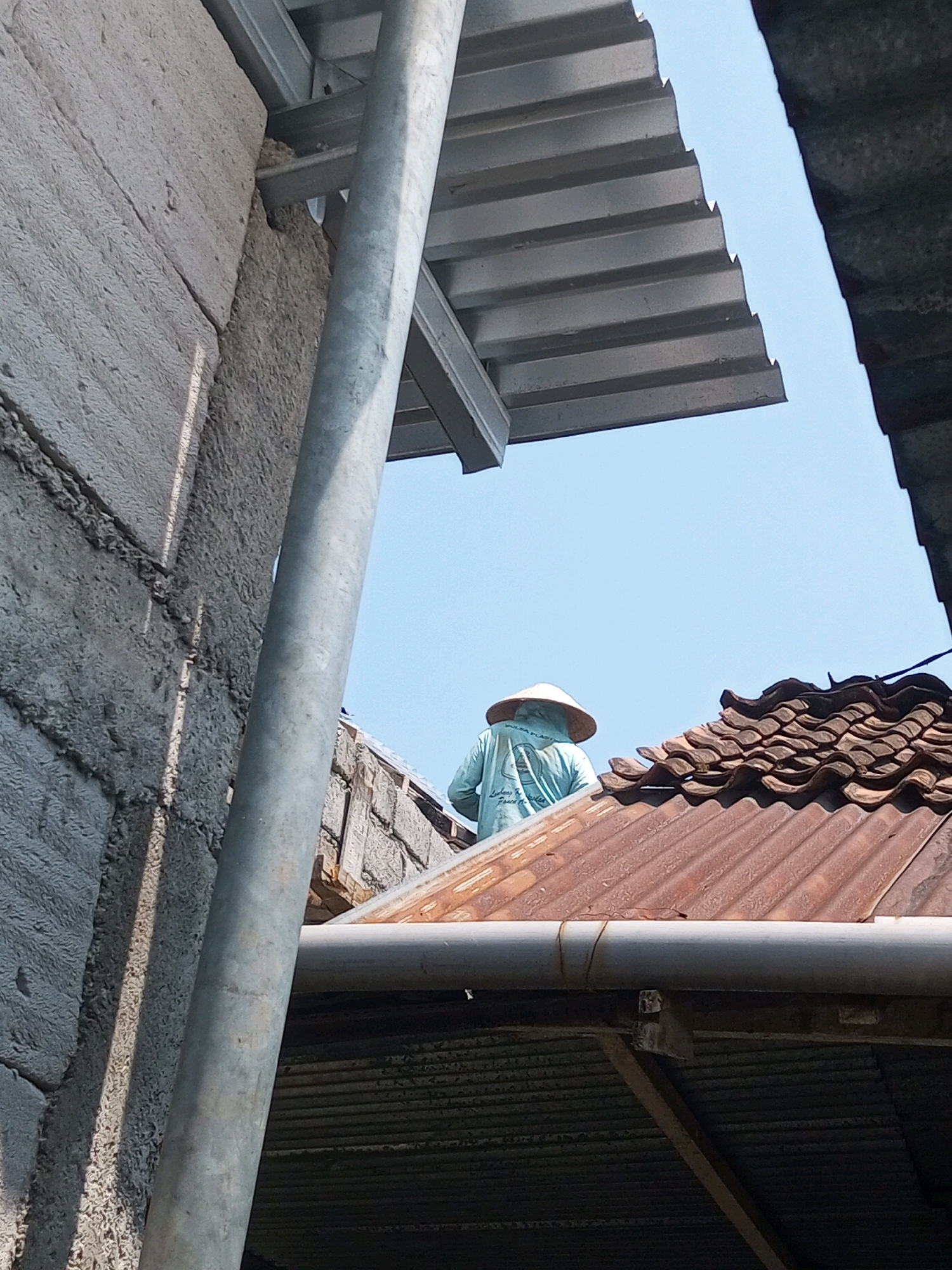 The image shows a construction site in Andongsili, Indonesia, with a worker in a blue shirt and conical hat on a terracotta tiled roof. The worker is partially obscured by a silver corrugated metal roof structure above. To the left, a rough, grey concrete wall is visible, with a vertical silver pipe running in front of it. The sky is clear and blue. The scene suggests ongoing construction or repair work.