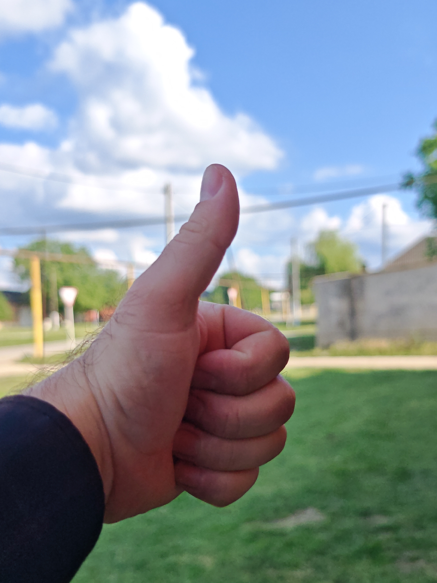A close-up, outdoor, eye-level shot shows a person's right hand with their thumb extended upwards in a "thumbs up" gesture. The hand is in the foreground and is partially in shadow from the sleeve of a dark garment worn on the arm. The background is blurred, showing a grassy area, a concrete wall, trees, and a bright blue sky with fluffy white clouds. The scene appears to be in an urban or suburban setting, possibly a park or residential area. The image was taken in Mayrtup, Russia.