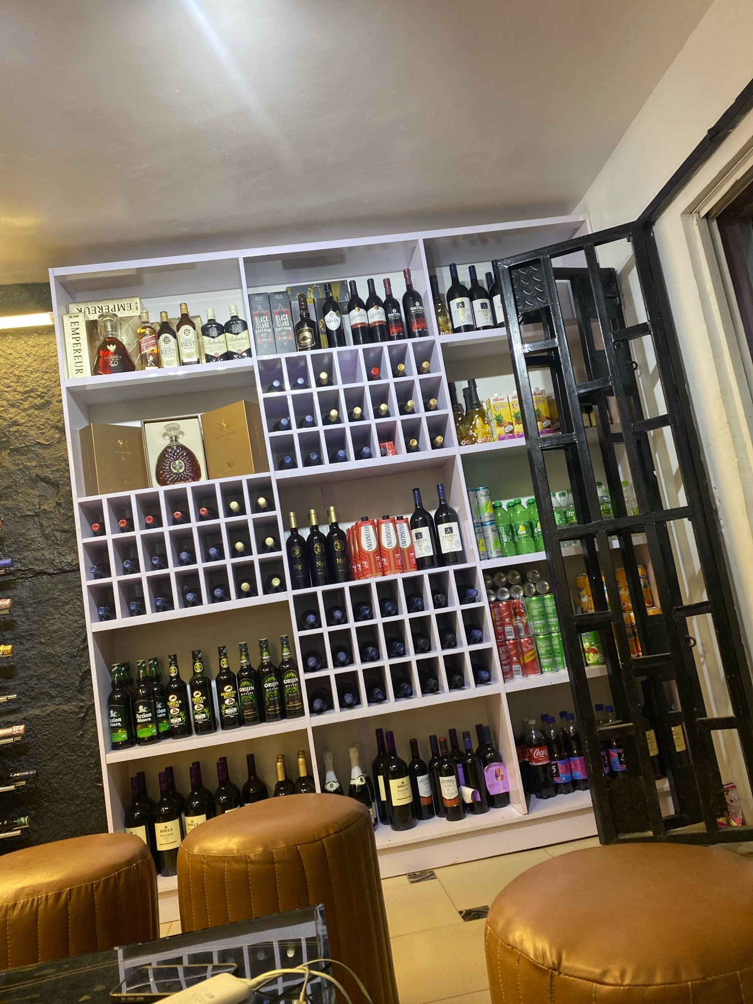 This is an indoor scene featuring a well-stocked liquor shelf in a store located in Jos, Nigeria. The shelf is white and filled with various bottles of alcohol, including wine bottles, liquor bottles, and some soft drinks and canned beverages. The bottles are arranged in cubbies and on shelves. In the foreground, there are two round, brown leather stools, suggesting a seating area for customers. A black metal grate or security gate is visible to the right of the shelves. The walls appear to be textured, with one section having a stone-like finish. The lighting is dim, creating a somewhat moody atmosphere.