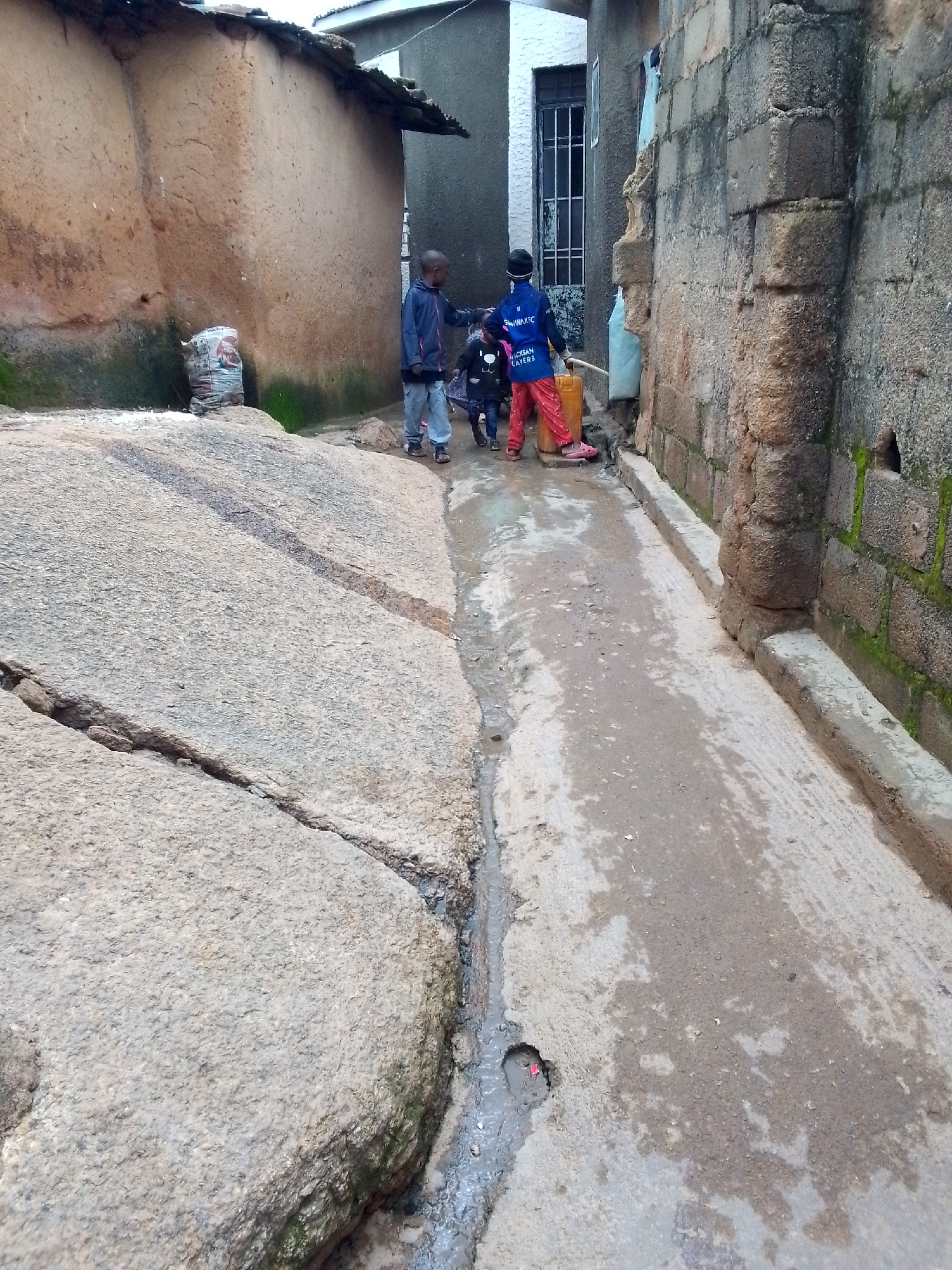 A street scene in Jos, Nigeria, features three children standing in a narrow, muddy alleyway between two buildings. A large, weathered rock formation occupies the left foreground, showing signs of erosion and cracks. To the left of the alley, a building constructed with mud plaster and a corrugated metal roof is visible, with a sack of goods placed near the base of the rock.
In the alley, the ground is damp and covered in mud, with a small puddle and a visible pothole. A concrete drain runs along the right side of the alley. The children are positioned near the center of the alley. The child on the left, facing away from the camera, wears a dark jacket and light-colored pants. The child in the center, also facing away, is dressed in a blue jacket with white lettering and red pants, holding a yellow jerrycan. The third child, to the right of the center child, appears to be wearing a dark outfit. A light blue plastic sheet is draped over a wall to the right.
The buildings flanking the alley are constructed of different materials. The one on the left is the mud-plastered structure, while the building on the right is made of rough-hewn stones, with visible moss growing in the crevices. A metal-barred window and door are present on this right-hand building. The overall impression is a somewhat neglected urban environment, likely in a residential area.