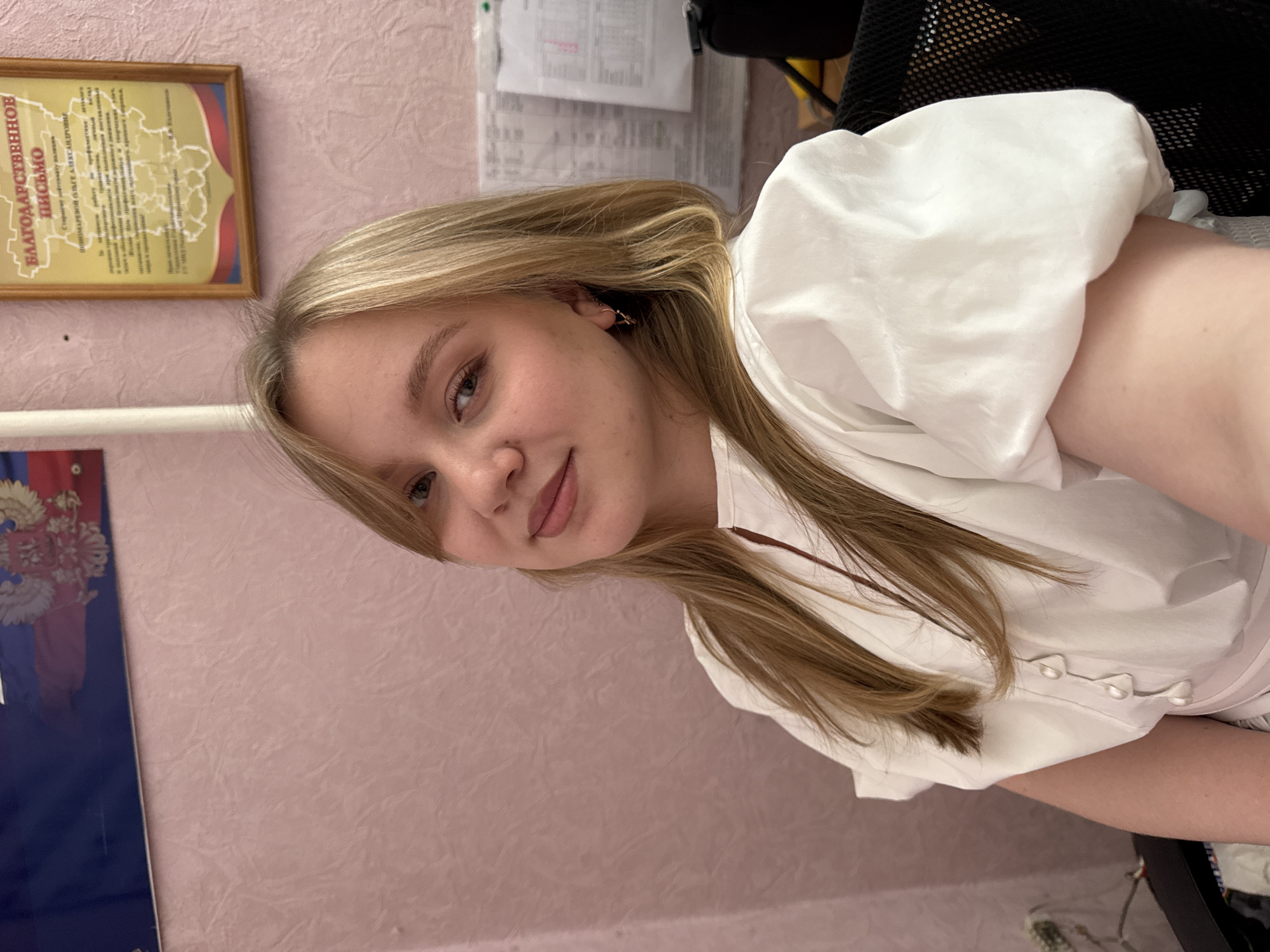 The media file is a portrait of a young woman with blonde hair, taken from a selfie perspective. She is wearing a white blouse with decorative buttons. Her face is turned towards the camera, and she is smiling slightly. In the background, there is a wall with textured pink wallpaper. To the left of the woman, a framed document with Russian text is visible. Below it, a portion of what appears to be a Russian flag is also visible. Further in the background, there are papers pinned to the wall, and part of a black office chair can be seen. The lighting suggests an indoor setting, likely during the daytime. The overall mood is casual and friendly.
Visible text on the framed document includes "БЛАГОДАРСТВЕННОЕ ПИСЬМО" which translates to "LETTER OF GRATITUDE". Below this, there is more text, but it is too small to be read clearly.