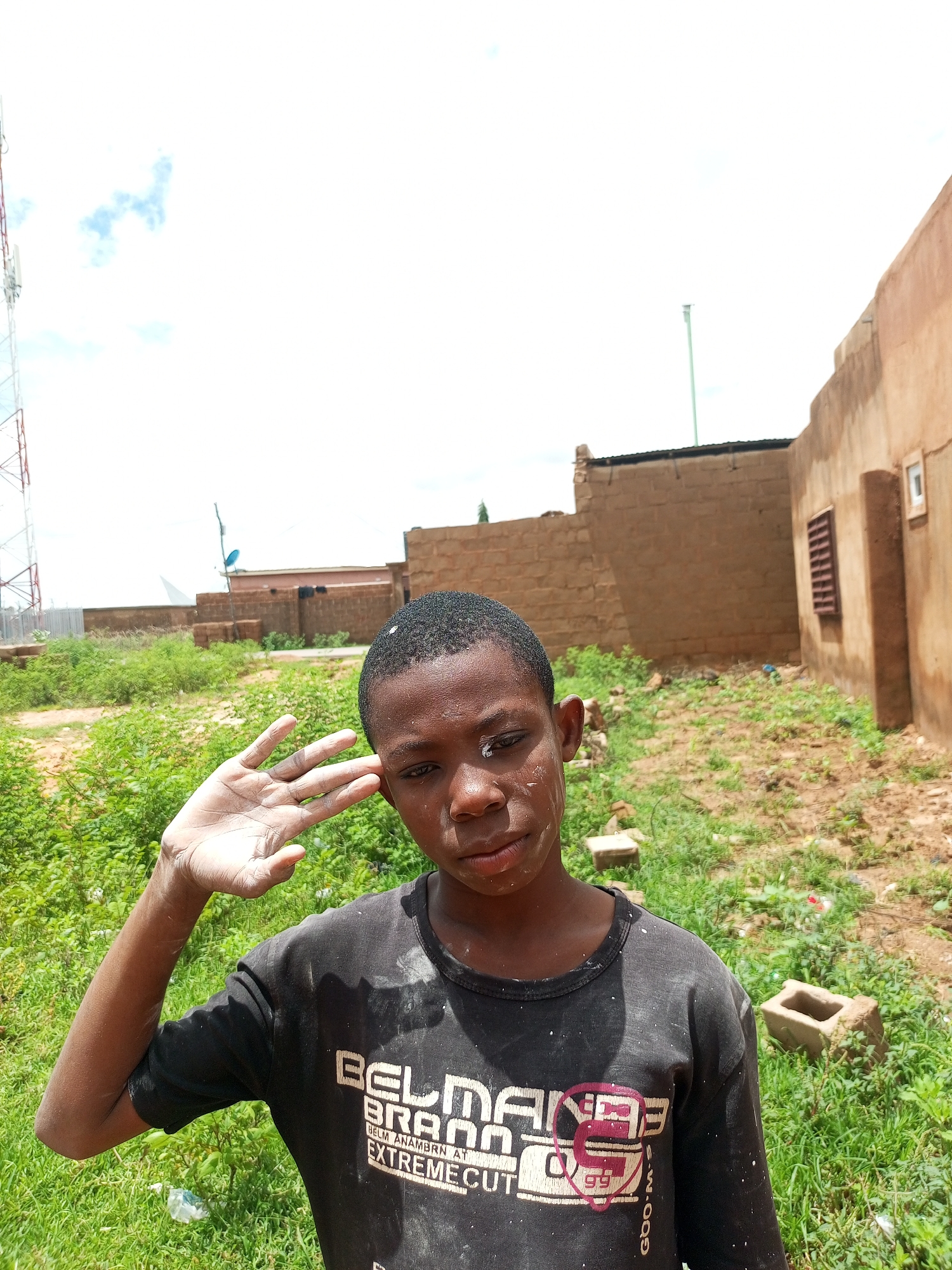 A young Black boy with dark hair, a light complexion, and visible dust on his face and shirt stands in a yard. He is wearing a black t-shirt with white text that reads "BELM BRAND EXTREME CUT". His right arm is raised, with his hand held to his forehead in what appears to be a salute. The background shows a dirt yard with patchy green grass, several concrete blocks, and a large building constructed of brick with a flat roof. To the left, a tall telecommunications tower is visible against a bright, cloudy sky. The setting appears to be an outdoor, undeveloped area within Tempure, Nigeria.