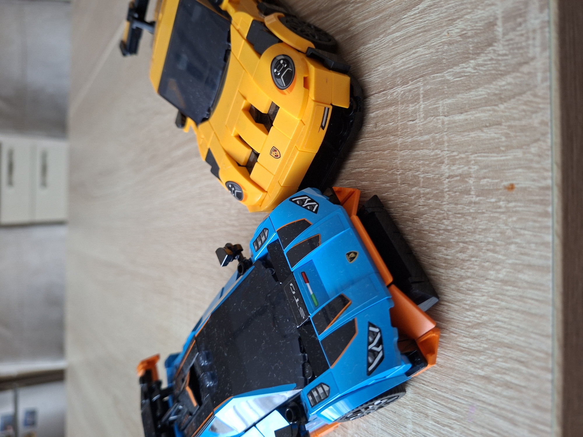 Two toy sports cars, a blue and orange Lamborghini and a yellow Porsche, are placed on a light-colored wooden table. The Lamborghini is on the left, and the Porsche is on the right. The cars are detailed replicas made of plastic bricks, likely LEGO. The background is blurred, showing what appears to be furniture. The scene is indoors, likely in a domestic setting, and no specific location in Baden-Baden, Germany, can be identified.