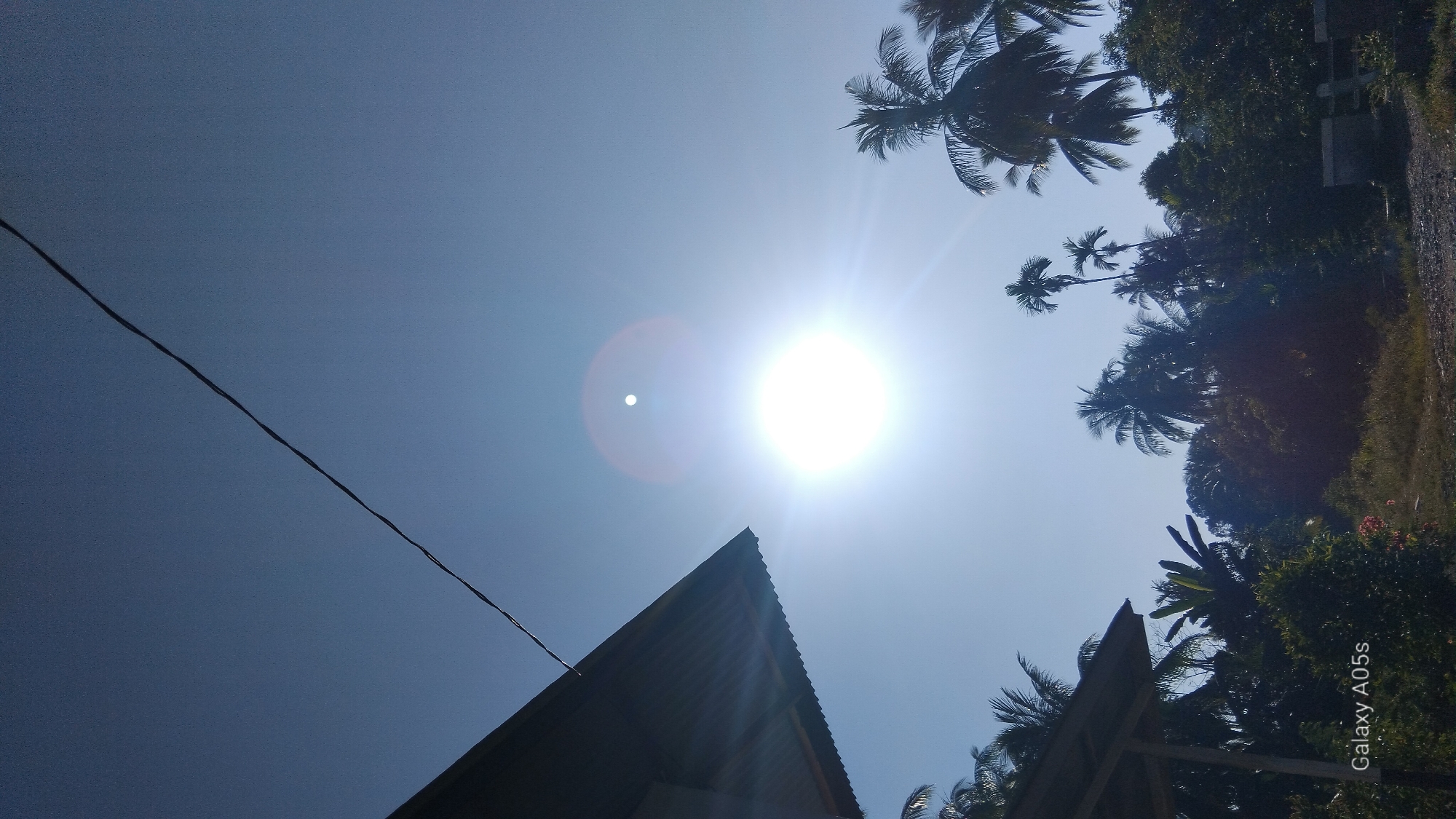 The image depicts a bright, sunny day with a clear blue sky. The sun is prominently featured, emitting strong rays and lens flare. In the foreground, a section of a corrugated metal roof extends from the left, casting a shadow. Below and to the right of the roof, lush green foliage and various trees, including palm trees, are visible. A thin, dark wire stretches diagonally across the upper portion of the sky. The bottom left corner of the image displays the text "Galaxy A05s". The scene appears to be in a tropical or subtropical environment, consistent with a location in Rambongpayong, Indonesia. There are no people or animals visible in the image. The overall impression is of a natural outdoor setting bathed in intense sunlight.