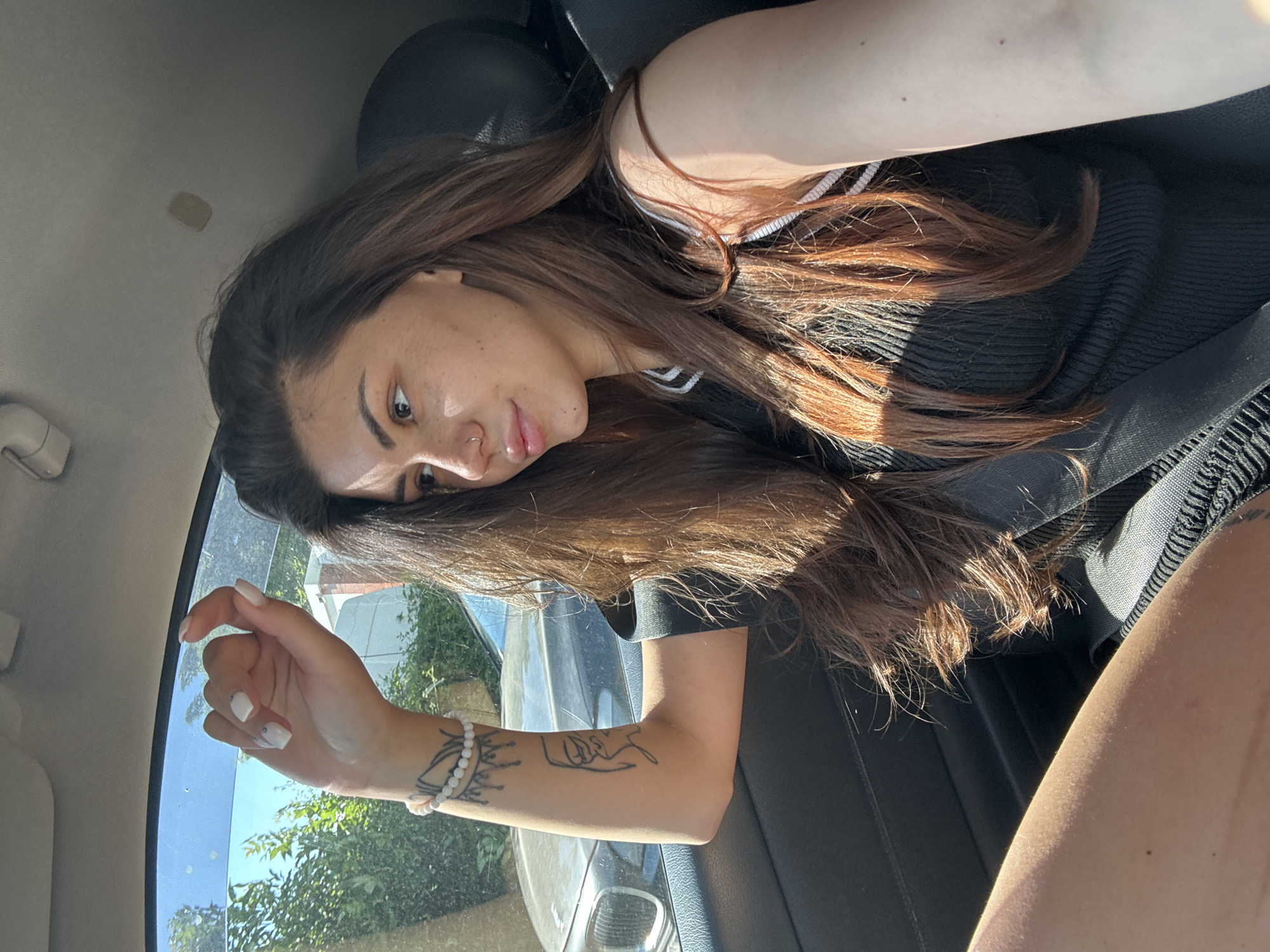 A young woman with long brown hair is sitting in the passenger seat of a car, looking out the window. She is wearing a black top. The sun is shining through the window, casting shadows on her face and hair. The car appears to be parked on a street in Sofia, Bulgaria, with trees and buildings visible in the background.