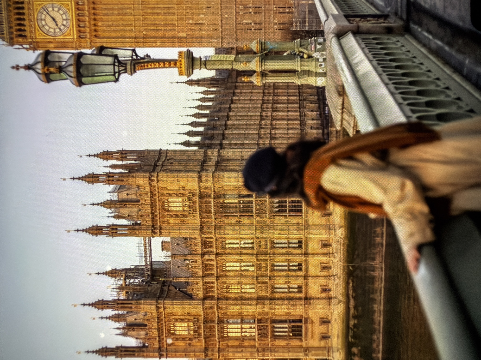 The image shows the Houses of Parliament and Big Ben in London, England, viewed from an elevated position, possibly a bridge. A person wearing a coat is visible to the right, leaning against a railing. The sky is overcast. A lamp post is visible in the foreground. The buildings appear to be constructed of light-colored stone, with many windows and architectural details.