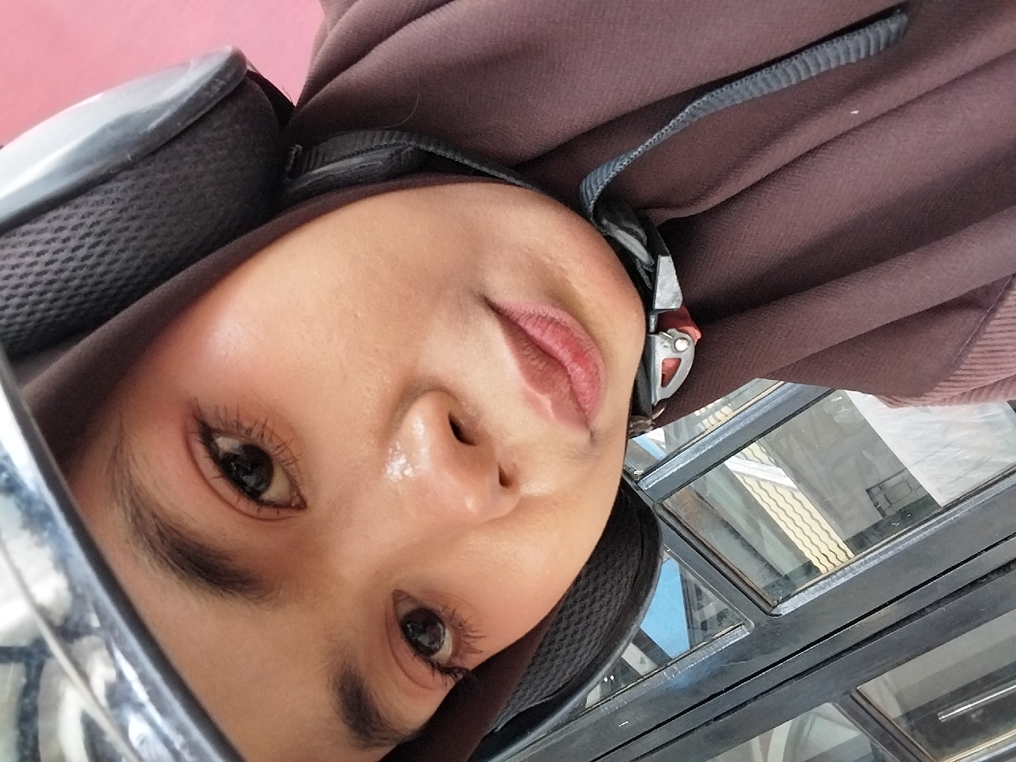 A young woman wearing a brown hijab and a helmet is captured in a close-up, selfie-style shot. Her face is turned slightly towards the camera, and she appears to be looking directly at it. Her makeup includes eyeshadow and lipstick. The background shows a window frame with glass panes, reflecting a building with a tiled roof. The woman's right shoulder and upper chest are visible, draped in her hijab. The image appears to be taken outdoors, likely in a semi-urban environment. The photo was taken at Jalan Trans Sulawesi, Tibojong, North Wanuakaka, Tibojong, Kab. Polewali Mandar, Sulawesi Barat, Indonesia.