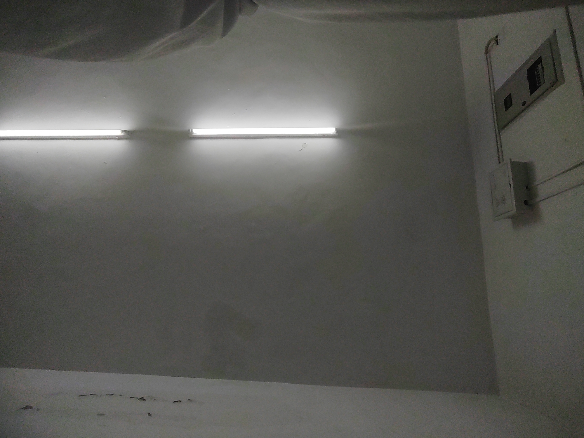The image shows an interior view of a room with white walls and ceiling. Two fluorescent light fixtures are mounted horizontally on the wall, providing illumination. On the right side of the wall, a grey electrical panel box is mounted. The floor is also visible and appears to be a light color with some discoloration and debris. The image is taken from a low angle, looking upwards towards the ceiling and the top portion of the walls. The lighting creates shadows on the wall and ceiling, suggesting the light sources are above and to the sides. There are no people or animals visible in the image. The scene appears to be an empty room, possibly a storage area or a utility space, located in the Chongqing High-tech Zone, China.