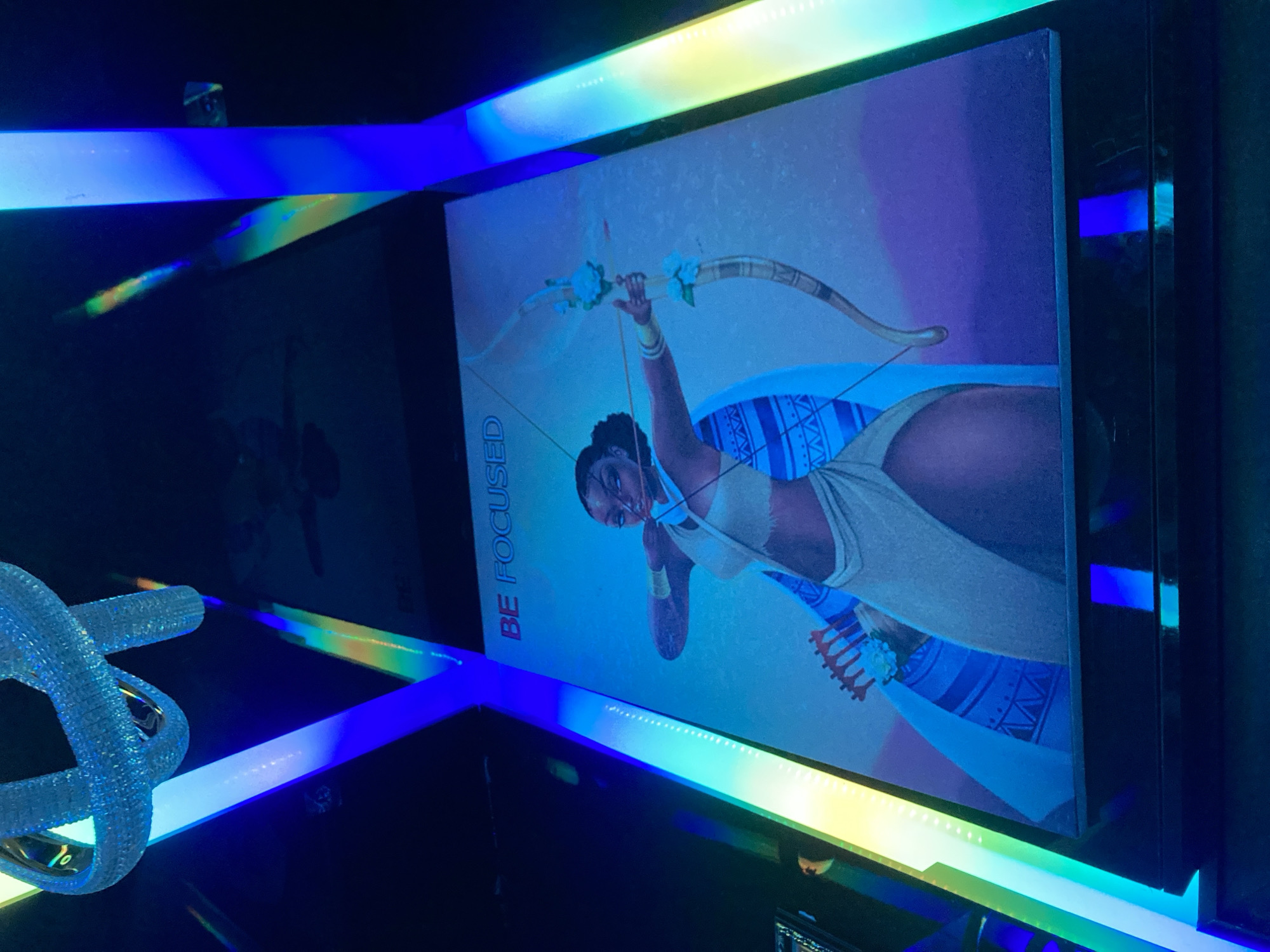 The image depicts a framed artwork featuring a woman with a bow and arrow, holding it as if to shoot. The artwork is illuminated by colorful LED lights in blue and green hues, creating a vibrant and dynamic atmosphere. The word "BE FOCUS" is printed in red letters at the top of the artwork. The artwork is displayed on a dark surface, possibly a wall or a frame. The overall setting appears to be an indoor space with modern lighting. The scene is shot from a slightly low angle, looking upwards towards the artwork and the lighting fixtures. The location in Abuja, Nigeria, is not specified within the image itself, but the context suggests it could be a gallery, a commercial space, or an event venue in the city.
