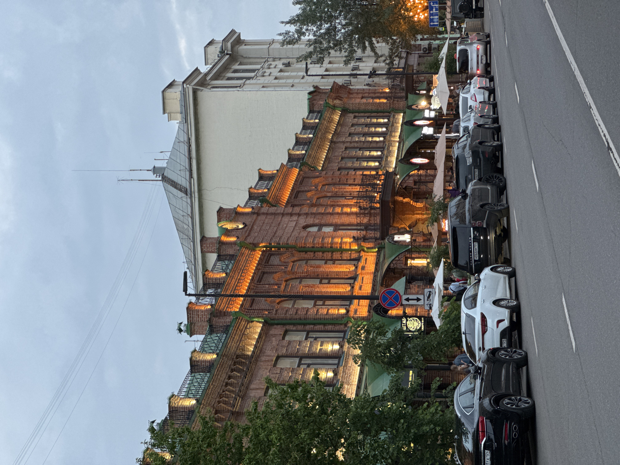 The image shows a street scene in Krasnoyarsk, Russia, with a row of buildings and parked cars. The architecture features brick facades with ornate detailing and illuminated windows, suggesting a historical or well-maintained urban area. A white Kia sedan is prominently parked in the foreground, alongside other vehicles including a black SUV and a silver hatchback. Several pedestrians are visible near the buildings, some seated at outdoor cafes. The sky is overcast, indicating either dusk or a cloudy day. Streetlights and the illuminated windows of the buildings cast a warm glow on the scene.
