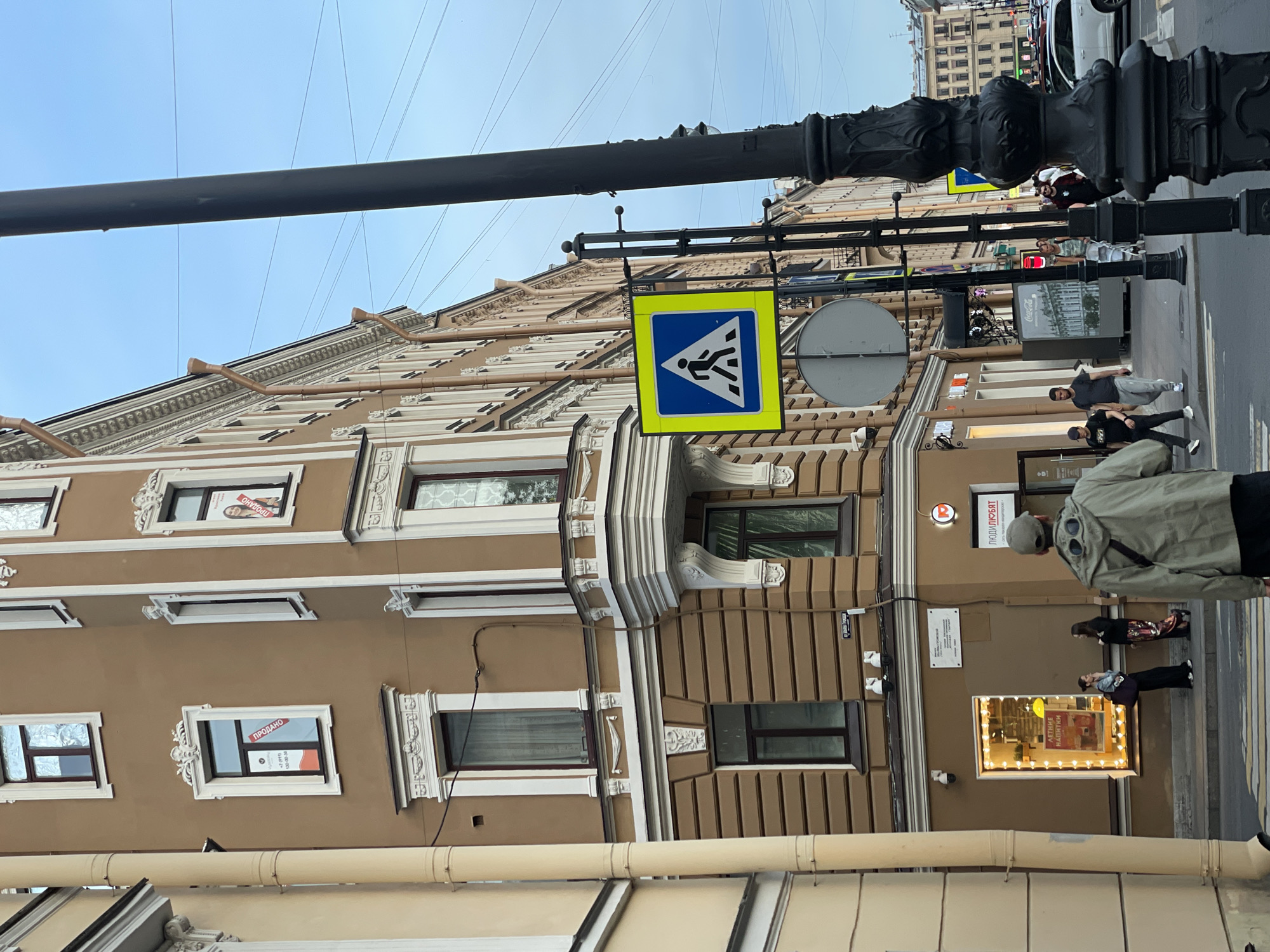 The image depicts a street scene at the intersection of Ulitsa L'va Tolstogo (Lev Tolstoy Street) and Ulitsa Professora Popova (Professor Popov Street) in Saint Petersburg, Russia. The dominant structure is a five-story corner building, painted a light brown or beige with elaborate white neoclassical architectural details, including decorative window frames, cornices, and a prominent bay window on the second and third floors.
Several windows on the upper floors display red "ПРОДАНО" (SOLD) signs in Russian. One such sign on the second floor includes a phone number +7 (911) 130-00-XX and a small logo. Another "SOLD" sign features a smiling woman's photograph. A light-colored, vertical drainpipe runs along the left side of the building. Two white security cameras are mounted on the building's facade above the ground-floor storefronts, and a third is located below the first-floor window.
The ground floor features commercial spaces. The leftmost visible shop has a brightly lit window display featuring a sign for "ЛЕТНИЕ НАПИТКИ" (Summer Drinks). To its right, another shop entrance is visible with a red circular logo and the text "ЛЮДИЛЮБЯТ" (People Love) above it.
In the foreground, a pedestrian, a man wearing a light green hooded jacket with distinctive goggle-like lenses on the hood, a baseball cap, and dark trousers, is walking away from the viewer towards a zebra crossing. He carries a dark shoulder bag. On the sidewalk outside the "Summer Drinks" shop, two women are standing; one is talking on a mobile phone, and the other observes the street.
A tall, black, ornate street light pole stands on the right side of the image, partially obscuring the background. To its left, a black pole supports a square yellow-and-blue pedestrian crossing sign (a white figure walking on a zebra crossing within a blue triangle). Below this is the back of a circular grey road sign. Further down the street, additional pedestrians are visible on the sidewalk, including two men, one in a black t-shirt and grey track pants, and another in a patterned shirt and shorts pushing a stroller.
The sky is a clear, light blue with several overhead utility wires crisscrossing the frame. In the background, other multi-story buildings line the street, and several cars, including a white SUV, are visible on the road.