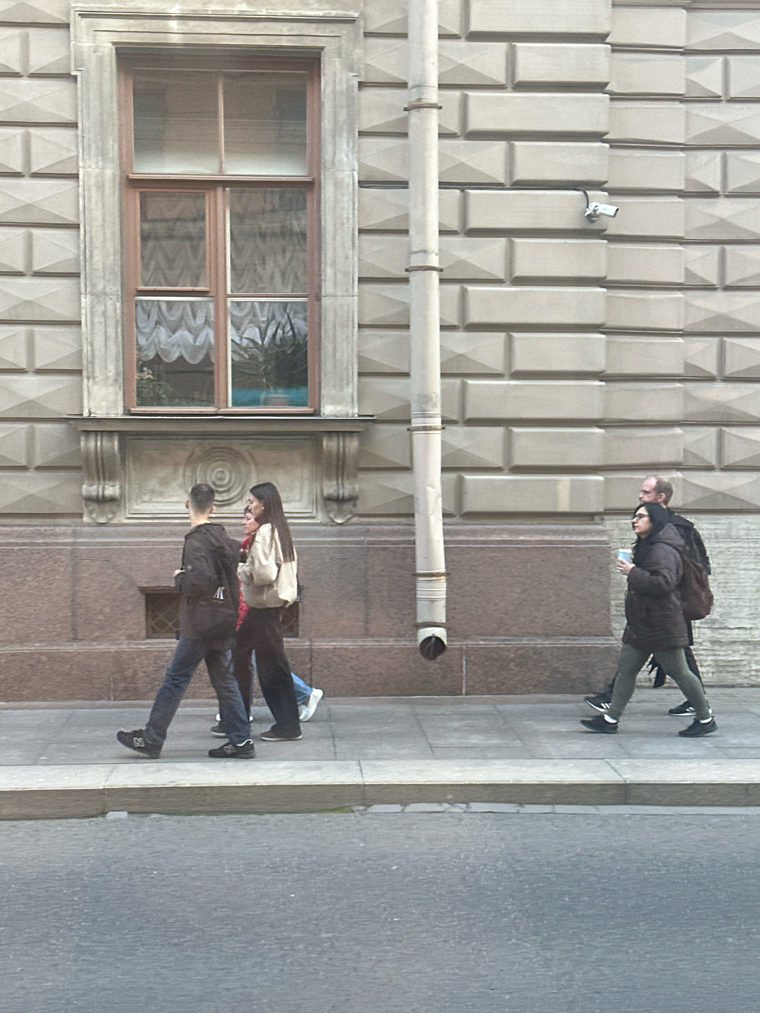 The image depicts a street scene in Saint Petersburg, Russia, with a focus on the facade of a building and several individuals walking on the sidewalk. The building features a large, multi-paned window with ornate trim, and its walls are constructed of textured stone blocks. A downspout runs vertically along the side of the building, and a security camera is visible mounted on the wall near the downspout.
In the foreground, four people are walking from left to right on the sidewalk. The first person, a male, is wearing a dark hooded jacket and blue jeans, carrying a backpack. To his right, a female is wearing a light-colored jacket and dark pants, also carrying a bag. Behind them, a third individual, partially obscured, is wearing blue jeans and dark shoes. Further to the right, another male is wearing a dark jacket, glasses, and dark pants, carrying a backpack, and holding a light-colored cup. A female is walking beside him, wearing a dark jacket, green leggings, and dark shoes, also carrying a backpack. The sidewalk is paved with concrete slabs, and a street curb separates it from the asphalt road. The overall lighting suggests an overcast day.