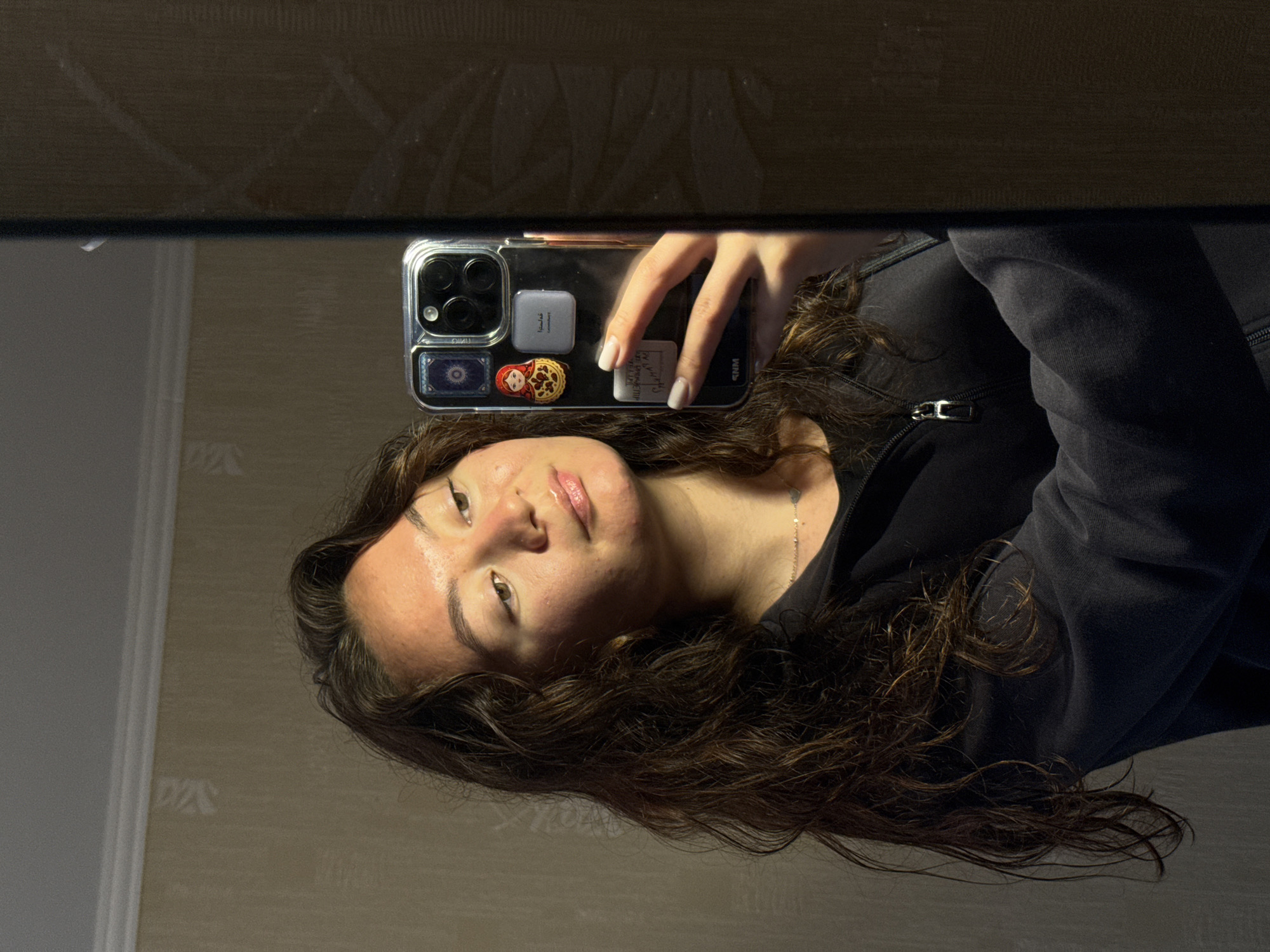 A young woman is taking a selfie in a mirror. The reflection shows the woman holding her phone to capture the image. The phone has a clear case decorated with several small items. She is wearing a dark hooded jacket. The environment appears to be a room with wallpaper and a door or piece of furniture visible in the mirror. There is no specific information in the image that indicates this photo was taken in Chaikovsky, Russia.