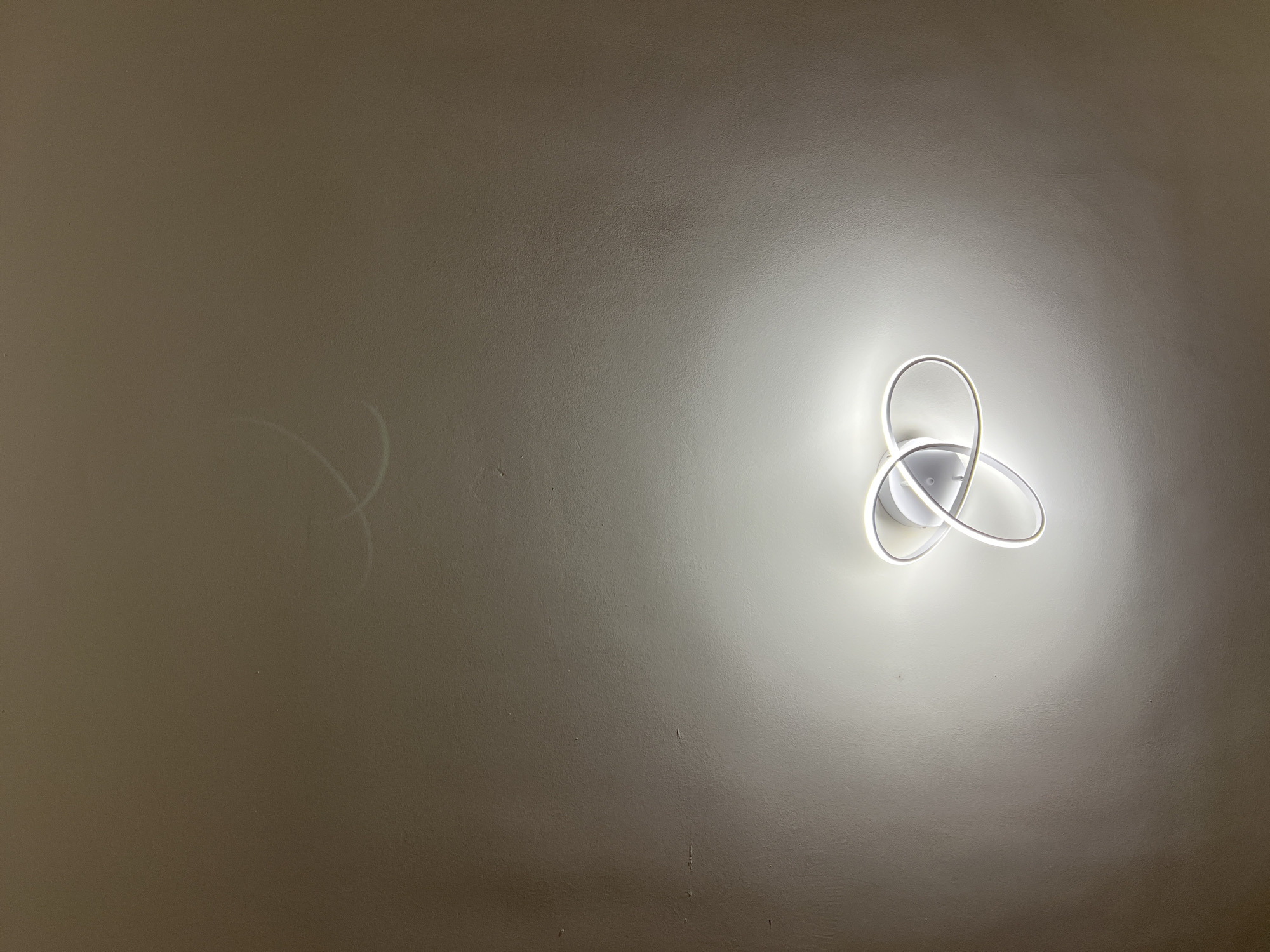 A modern, white ceiling light fixture with intertwined, circular LED elements illuminates a textured, neutral-colored ceiling. Faint, light-colored marks, possibly water stains or scuffs, are visible on the ceiling above the light fixture. The image is taken from a low angle, looking upwards. This scene is not discernable as being in Verona, Italy.
