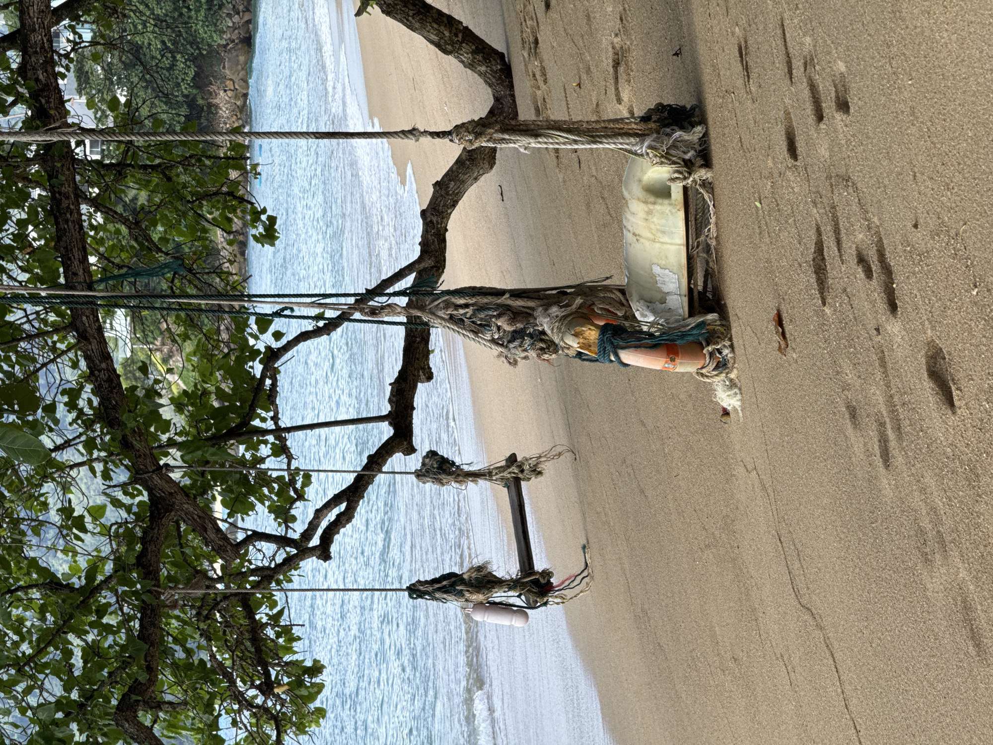 This image depicts a tranquil beach scene in Kamala, Thailand, under a tree with lush green foliage. Two swings are suspended from the tree's branches. The swing on the left is made of a weathered wooden plank and is adorned with a cylindrical white buoy. The swing on the right features a more makeshift seat, possibly a repurposed plastic container, and is surrounded by frayed ropes. Both swings show signs of wear and tear, with ropes appearing old and weathered. The sand on the beach is golden brown, with several footprints visible, suggesting recent human activity. The ocean waves gently lap the shore, creating a serene atmosphere. In the background, a rocky coastline and greenery can be seen, adding to the natural beauty of the location.