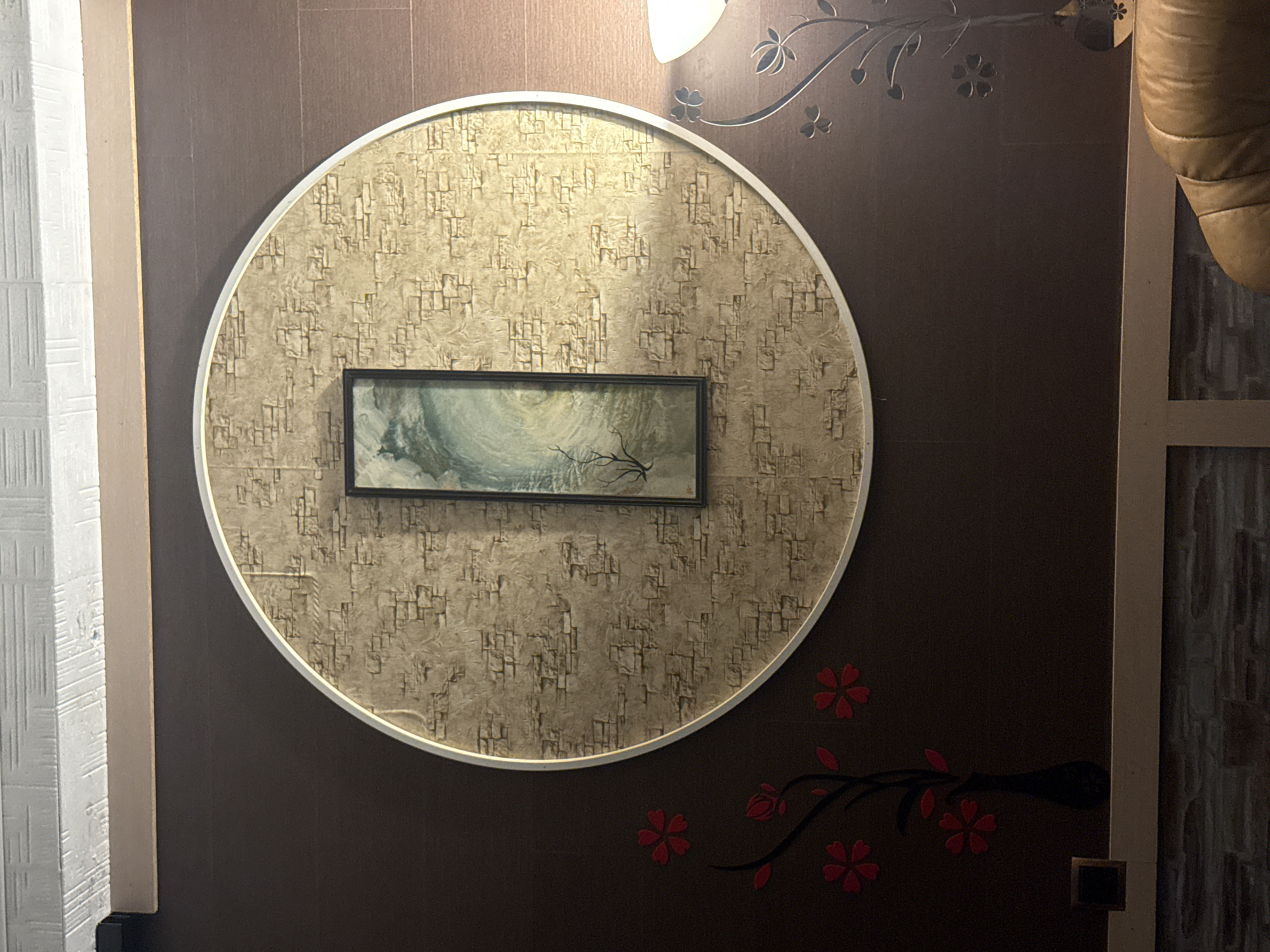 The image shows a close-up of a wall in a room, with a large circular decorative element featuring a textured, brick-like pattern. A narrow, rectangular painting of a swirling green and blue landscape with a bare tree is centered within the circle. To the left of the circle, on the dark brown wall, are red floral decals. Below the circular decoration, a thin shelf or mantelpiece is visible, with a small dark vase on the left and a corner of a brown leather armchair on the right. A lamp with a white shade is partially visible on the far right. The ceiling above the wall has a textured white finish. The scene appears to be an interior decoration. The location provided, Galinė, Lithuania, is a town, and this is likely a residential or commercial space within that town. There are no people or actions depicted.