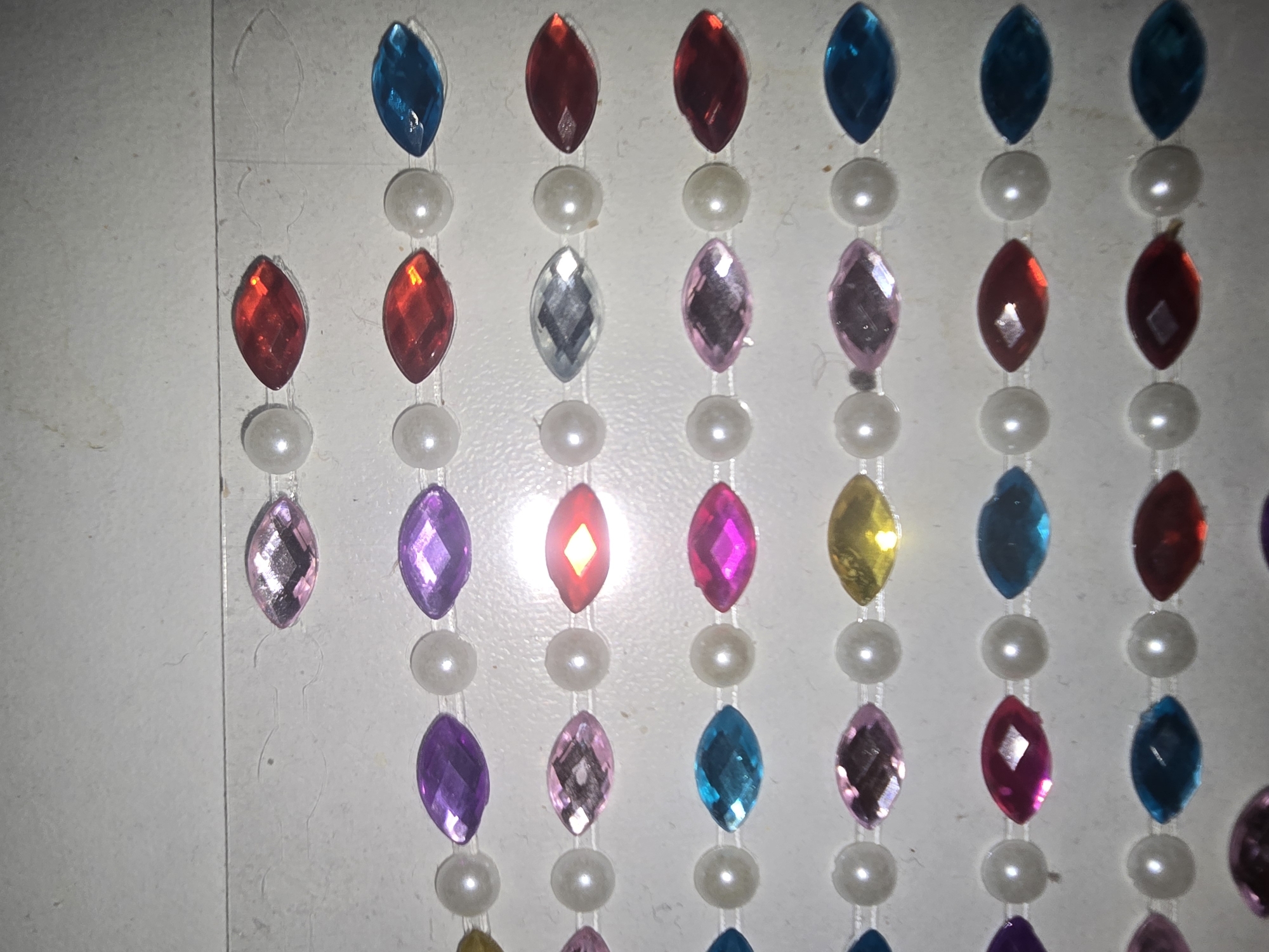 The image displays strings of colorful, marquise-shaped plastic jewels interspersed with small, white faux pearls. These strings are arranged in horizontal rows against a white, textured background. The jewels are primarily in shades of pink, purple, red, blue, and yellow. The lighting in the image creates glare on some of the jewels and pearls. There is no indication of location within the city of Ulan-Ude, Russia, as the image is a close-up of craft materials.
