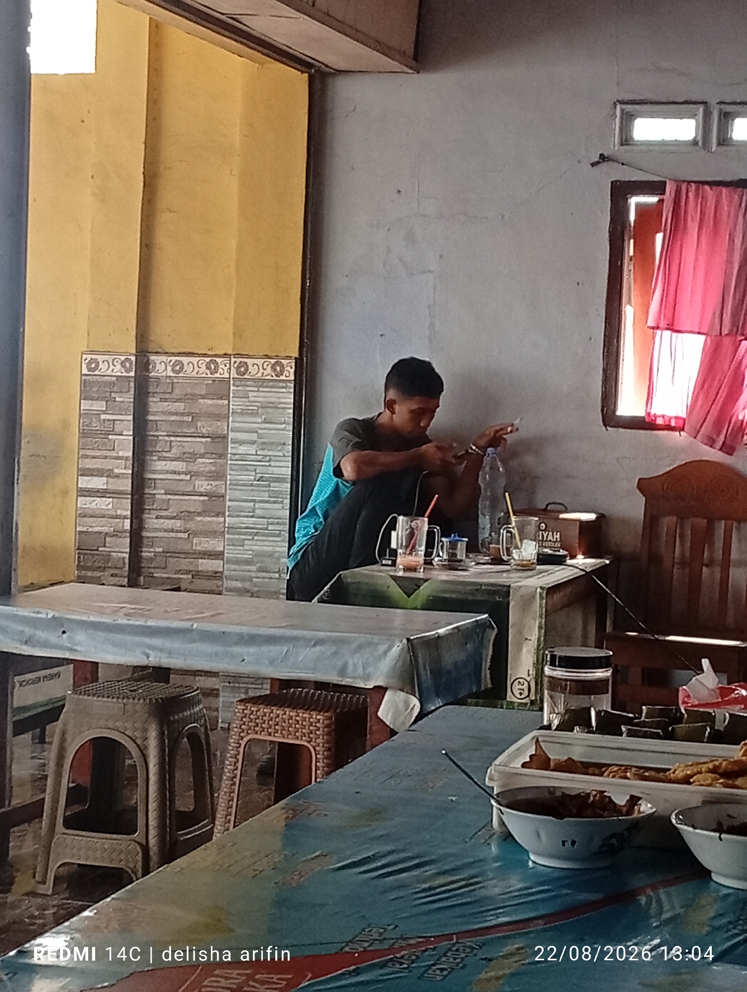 The image depicts an indoor scene, likely a food stall or eatery, in Gambir Kuning, Indonesia. A young man, appearing to be in his late teens or early twenties, is seated at a table, facing away from the camera. He is wearing a dark t-shirt and black pants, and is holding a cigarette in his right hand, with a mobile phone in his left. He is looking down at his phone. On the table in front of him are several items, including a bottle of water, cups with drinks, and possibly some food.
The environment suggests a simple establishment. The walls are plastered and painted a light gray, with a section of yellow-painted wall visible to the left, adorned with decorative tiles at the base. A window with red curtains is visible on the right wall. In the foreground, another table is covered with a blue and white patterned cloth, with several bowls of food on it, including what appears to be rice and fried items. Two plastic stools are positioned under the table in the middle ground. The lighting suggests it is daytime, with natural light entering from the window. The overall impression is that of a casual, everyday setting in a local Indonesian community. The timestamp in the lower right corner indicates the photo was taken on August 22, 2026, at 13:04.