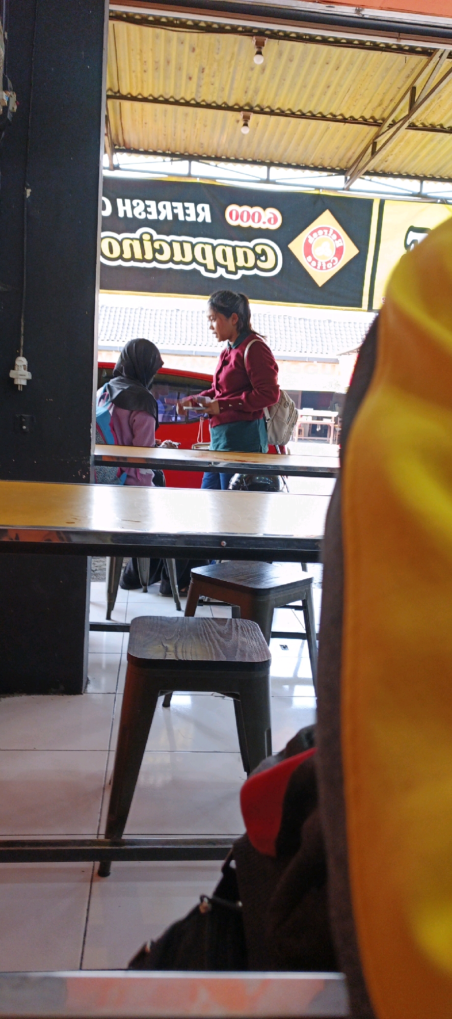 Two individuals are seen at a cafe table in Sidoarum, Indonesia. A woman wearing a maroon sweater and a green shirt is standing and facing a second individual who is seated and wearing a hijab and a purple jacket. The woman in the maroon sweater is holding a book or magazine. The cafe has a yellow corrugated ceiling. Tables and chairs are visible in the foreground.