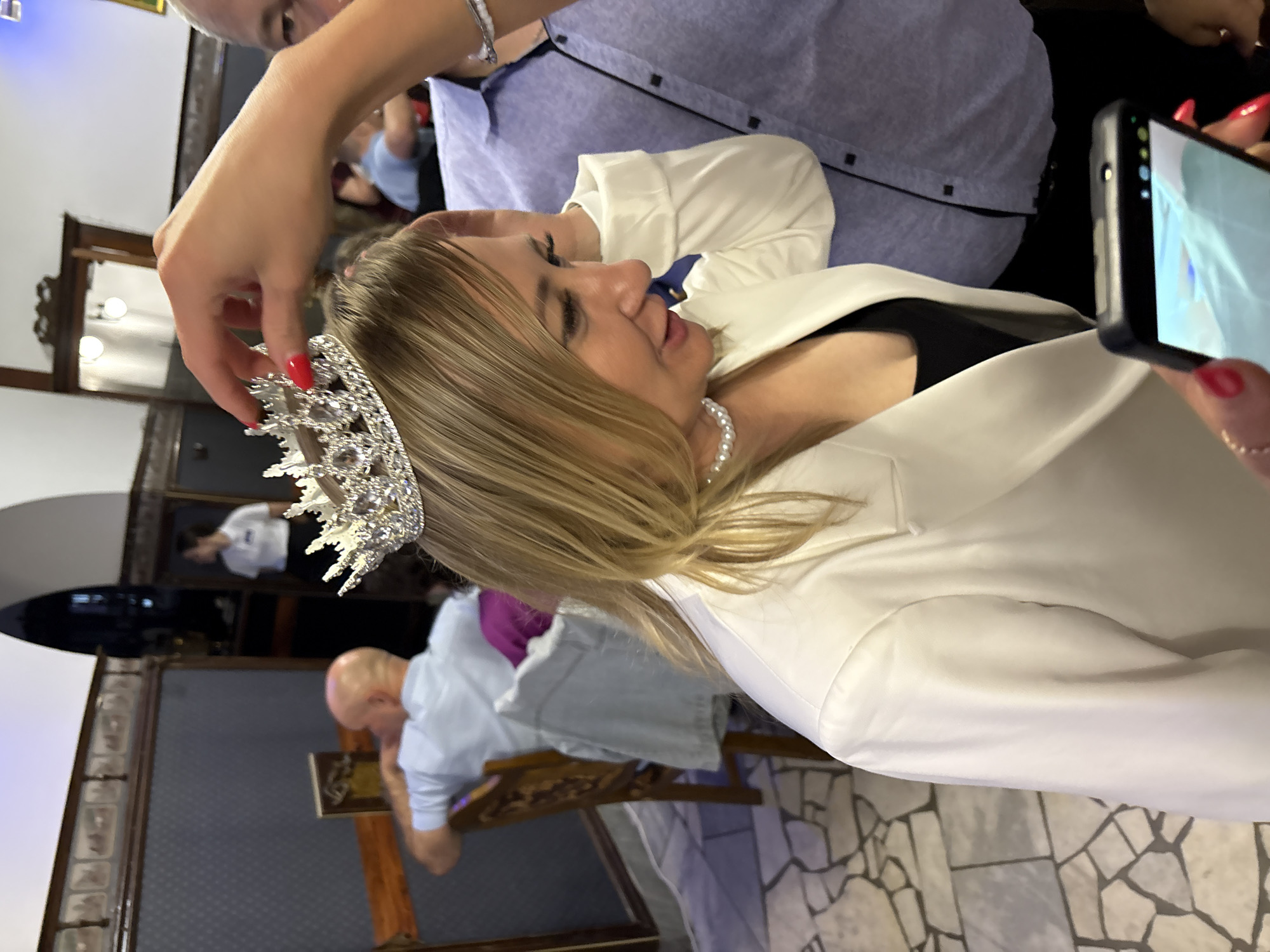 In Zavod Mikhailovskii, Russia, a woman with blonde hair is having a tiara placed on her head by a person whose hand is visible in the frame. The woman is wearing a white blazer over a black top and a pearl necklace. She has red nail polish. A man in a blue shirt is partially visible on the right side of the frame, and another man with a bald head is seated in the background on the left. The environment appears to be an indoor venue with tiled flooring and decorative wall panels.