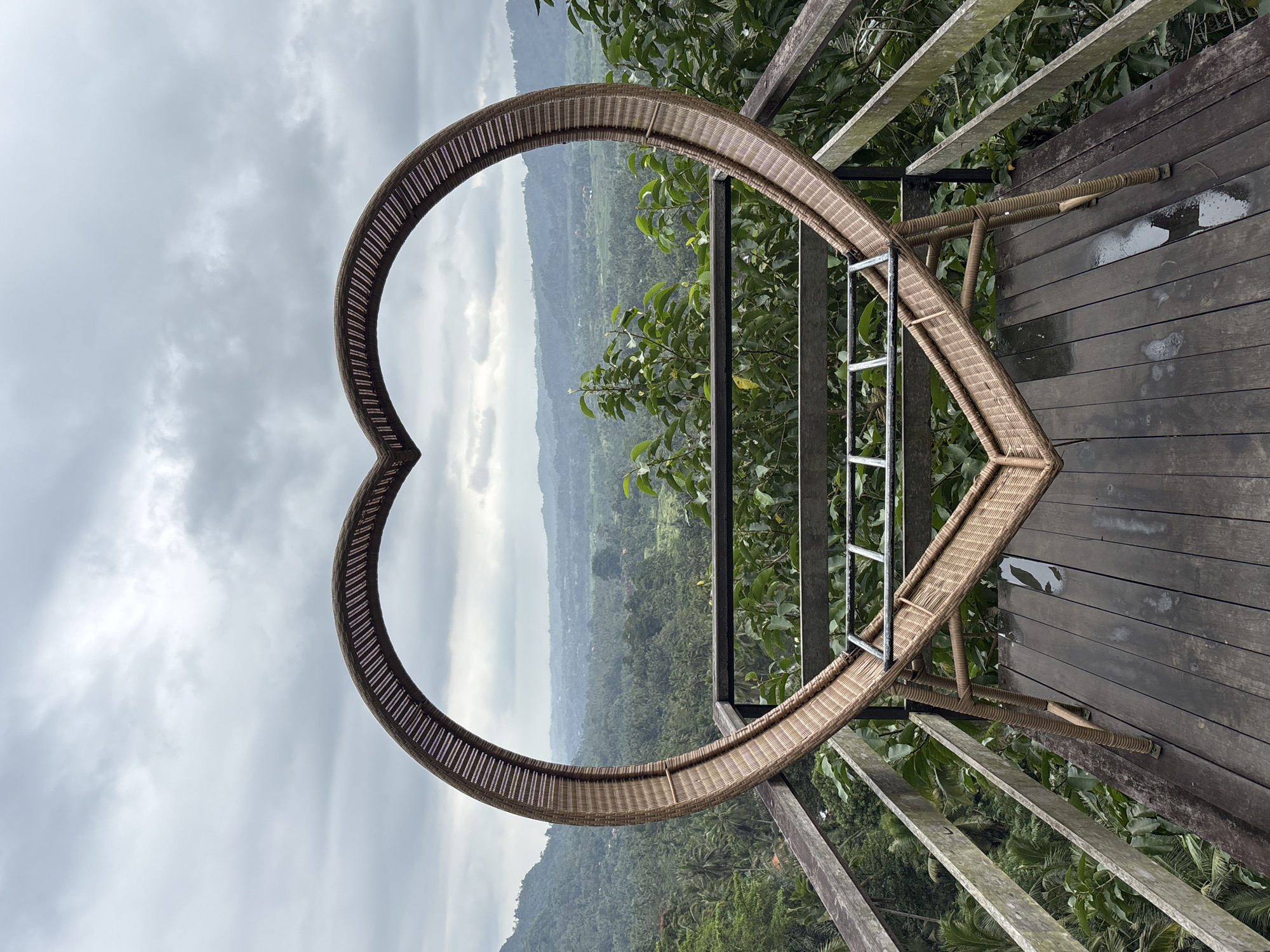 A heart-shaped rattan frame stands on a wooden deck overlooking a lush, verdant valley. The frame is woven with intricate patterns, and its open interior frames a panoramic view of rolling hills covered in dense tropical foliage. The sky above is overcast with dramatic clouds. A wooden railing borders the deck, providing a safety barrier. The deck itself is constructed from weathered wooden planks, some of which appear damp, suggesting recent rain. In the background, the valley stretches out, revealing more trees, scattered buildings, and mist-shrouded mountains in the distance. The scene evokes a sense of tranquility and natural beauty. This image is located in Sidemen, Indonesia, a region known for its stunning rice terraces and natural landscapes.