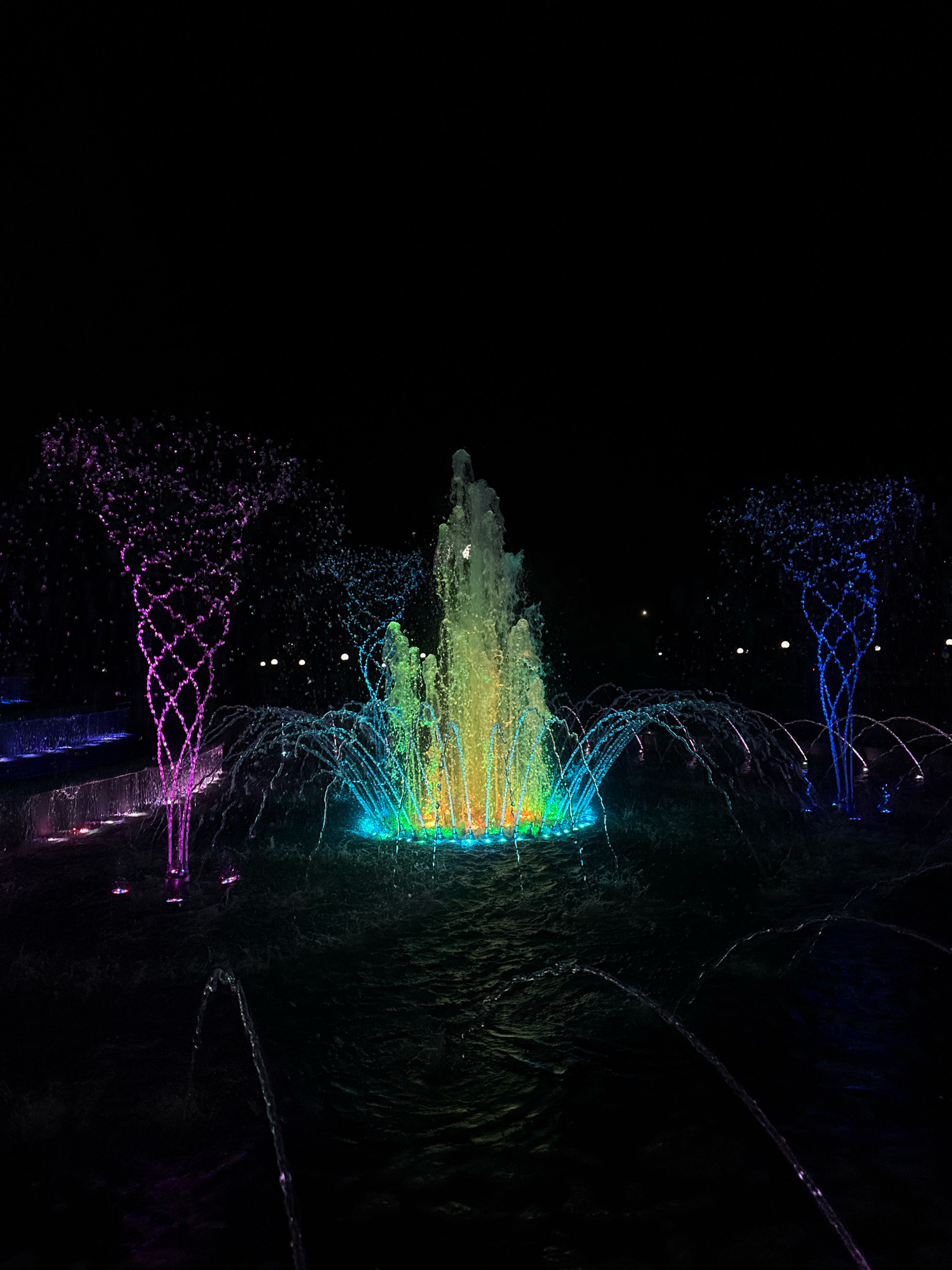 The image depicts a nighttime view of a decorative fountain in Mikashevichy, Belarus. The fountain features multiple jets of water illuminated by colorful lights, creating a vibrant spectacle against the dark background. In the center, a large jet of water is illuminated with a spectrum of colors, transitioning from green and yellow at the base to blue and purple at the top. To the left and right of the central jet, two smaller fountains are also lit, with the one on the left displaying purple hues and the one on the right showing blue. The water from these fountains forms intricate patterns as it falls back into the pool. The surrounding area is dark, with only a few faint lights visible in the distance, suggesting an urban setting. The surface of the water in the pool is rippled, reflecting the colorful lights and adding to the dynamic visual effect. There are no people or animals visible in the image. The scene is designed for aesthetic display, likely in a public park or plaza.