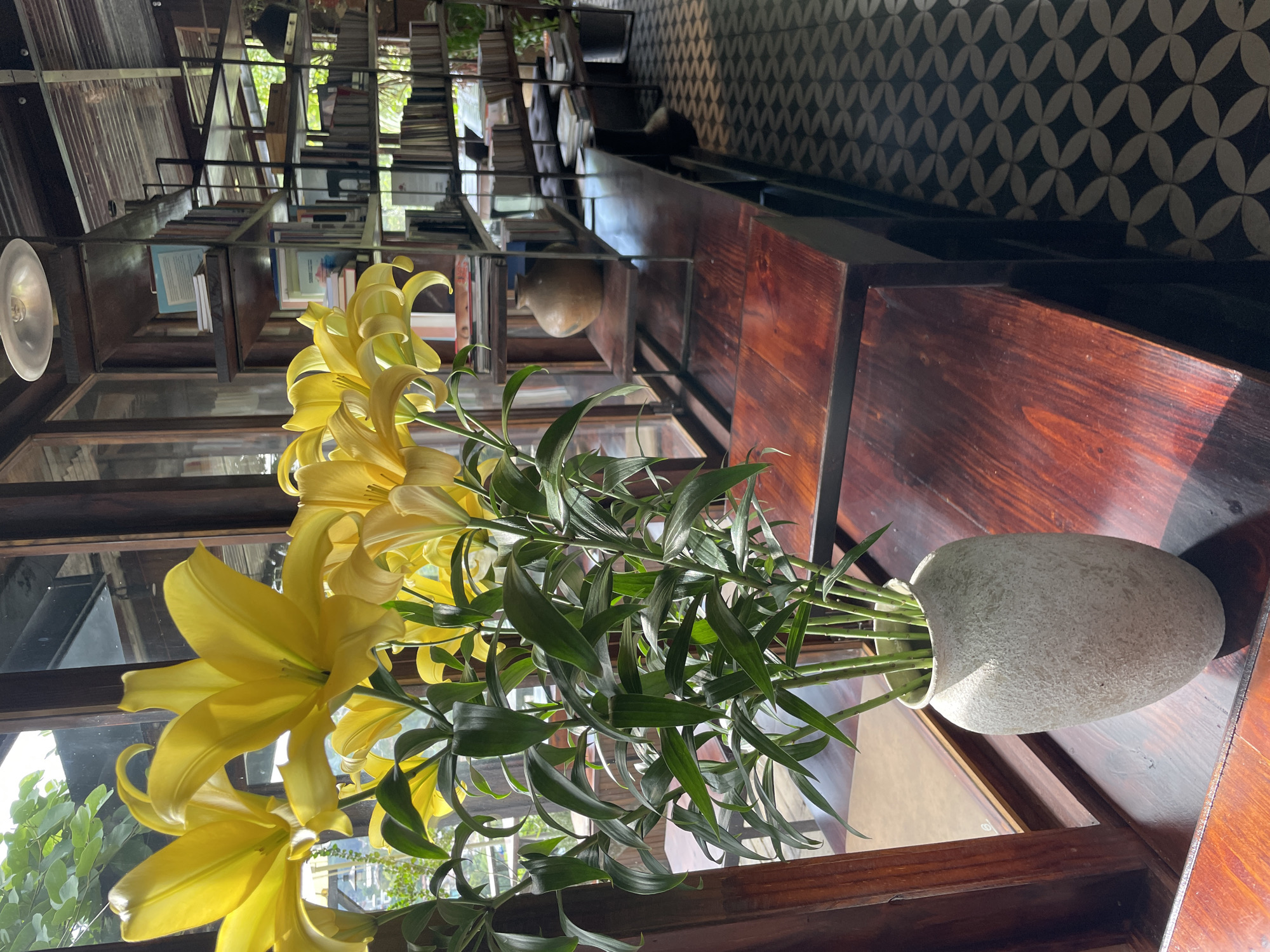 A close-up, eye-level shot captures a bouquet of vibrant yellow lilies in a textured, light-colored vase, placed on a dark, polished wooden surface. The lilies, with their prominent stamens and elegantly curved petals, dominate the left side of the frame. Behind them, a tall, dark wooden bookshelf filled with books stretches towards the right, with a single round, earthenware vase on a lower shelf. The environment suggests an interior space, possibly a cafe or library, with a window to the left allowing natural light to illuminate the scene. The floor features a repeating geometric pattern in black and white. The overall ambiance is one of quiet elegance and natural beauty. This scene is located at Da Lat Train Station, Da Lat City, Lam Dong Province, Vietnam.
