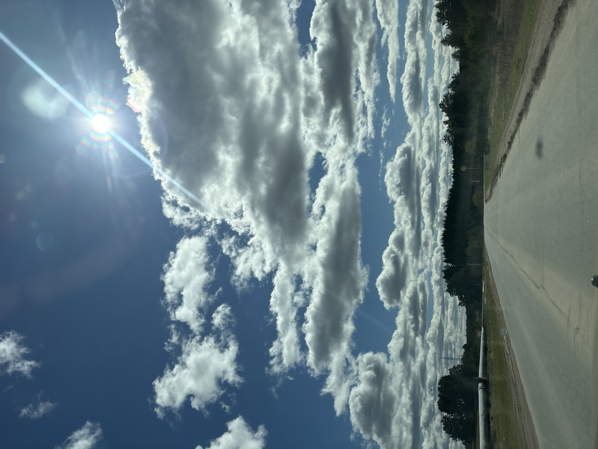 The image depicts a sunny sky with cumulus clouds over a road lined with trees. The sun is visible in the top left corner, causing lens flare. A gray, asphalt road extends into the distance on the right side of the frame. Trees form a dark green line along the road's edge. A pipe is running from the lower part of the image along the road. The image was taken outdoors in daylight. The location is near Прикамский, Russia.