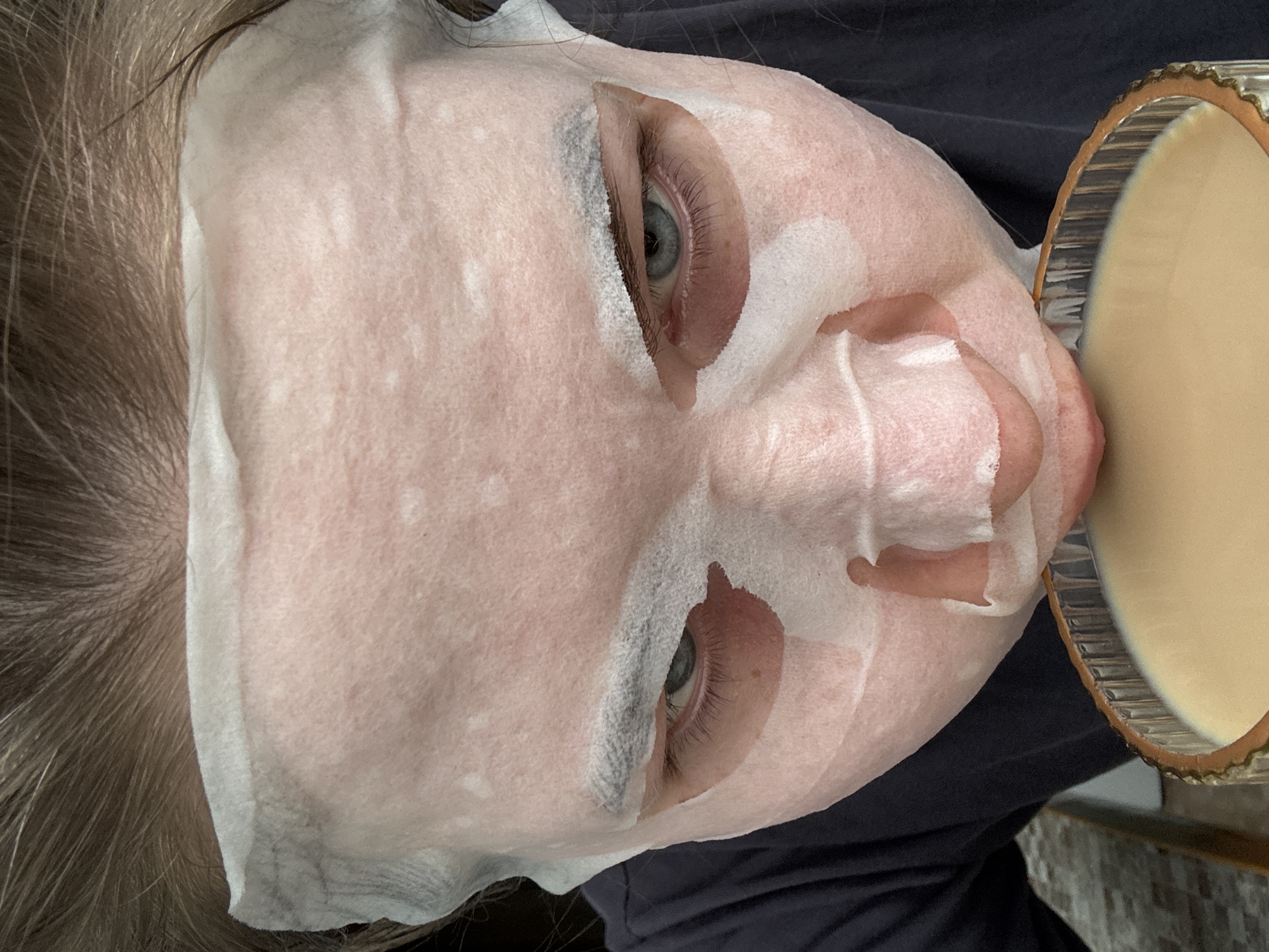 A close-up, slightly rotated image shows a person with a white sheet face mask. The mask has openings around the eyes and nostrils. The person's left eye (from the viewer's perspective) is visible, with a blue iris. Light brown hair is partially visible on the left side of the frame. A round object with a light tan interior and textured bronze-colored rim is on the right side of the frame. A dark-colored garment is visible beneath the person's head. The location is not evident in the image. There is no information in this image to determine the location in the city of Chaikovsky, Russia.