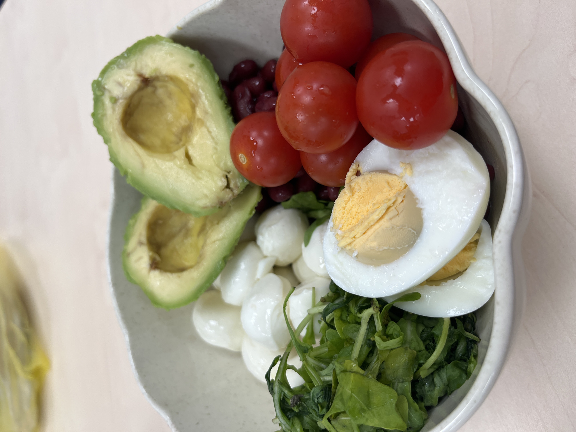 A close-up, eye-level, top-down shot captures a healthy salad in a light-colored, irregularly shaped bowl. The bowl contains a variety of ingredients: two halves of ripe avocado, a pile of small, white mozzarella balls, a generous portion of fresh green spinach leaves, several bright red cherry tomatoes, some red kidney beans, and one hard-boiled egg, cut in half, revealing a yellow yolk and white. The salad is placed on a light-colored, wooden surface. There are no people or specific city landmarks visible to determine a location in Plovdiv, Bulgaria or any other city. The focus is solely on the food.