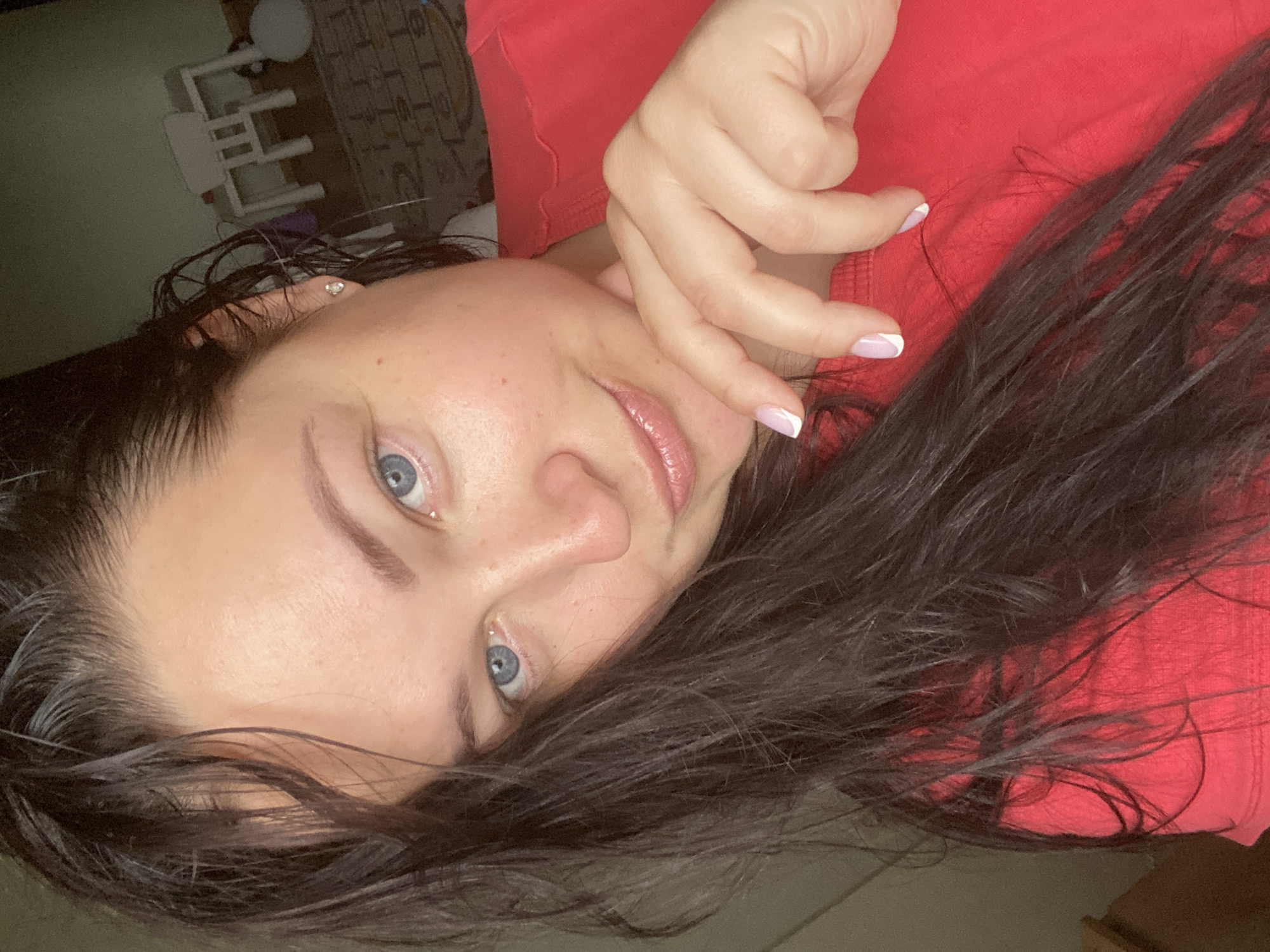 A woman with blue eyes and dark hair lies with her head turned towards the camera. She is wearing a red top. Her hand is near her face, and her nails have white tips. The background shows a glimpse of a room with white furniture. The location is not discernible as being in Sochi, Russia from the provided image.