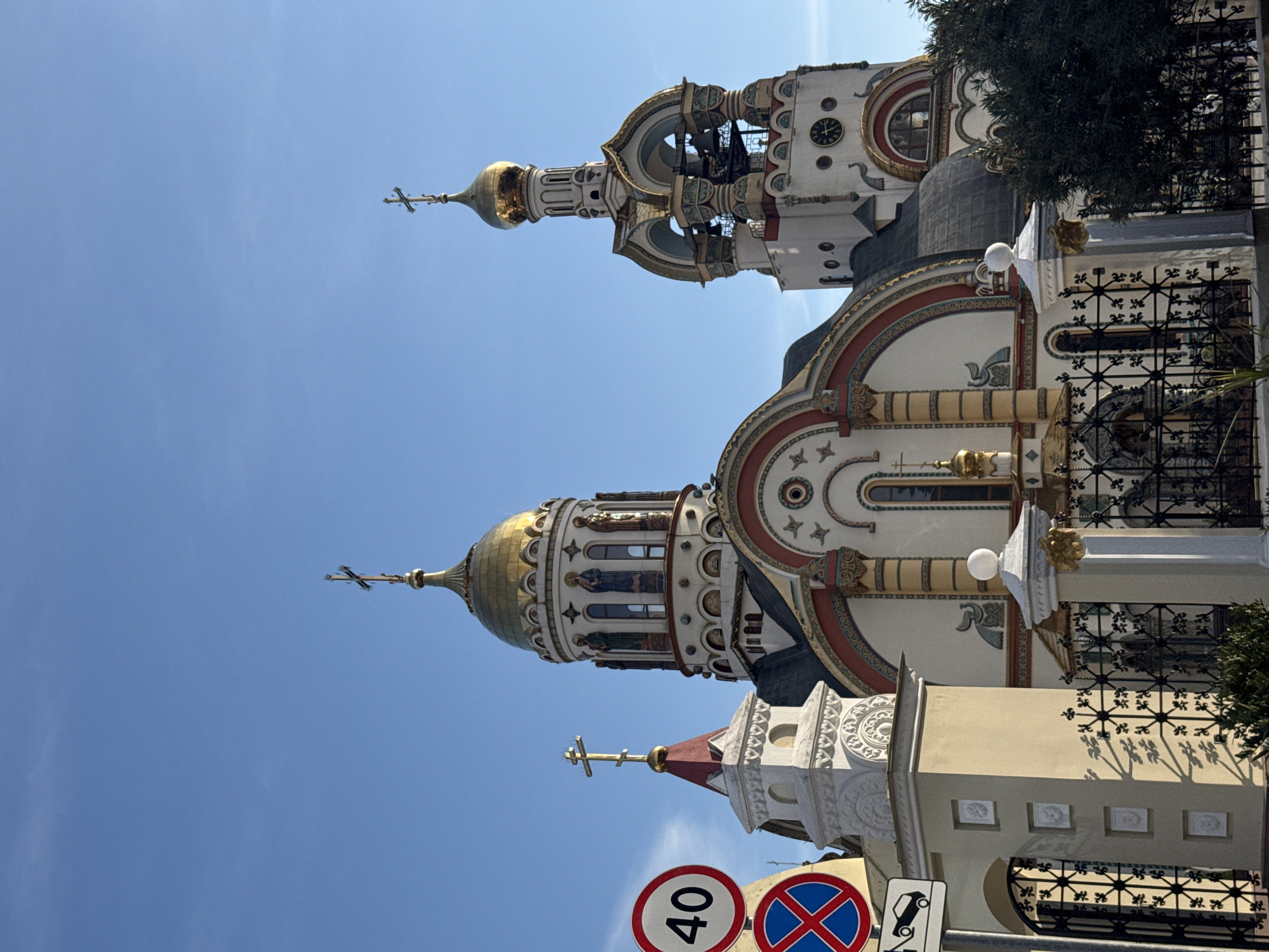 The image depicts the Holy Trinity Church in Adler, a district of Sochi, Russia. The church is adorned with golden domes and crosses. It features intricate architectural details, including decorative columns, arched windows, and ornate embellishments. The facade is primarily white with red and gold accents. In the foreground, traffic signs are visible. A blue sky serves as the backdrop. A tree is seen on the right.