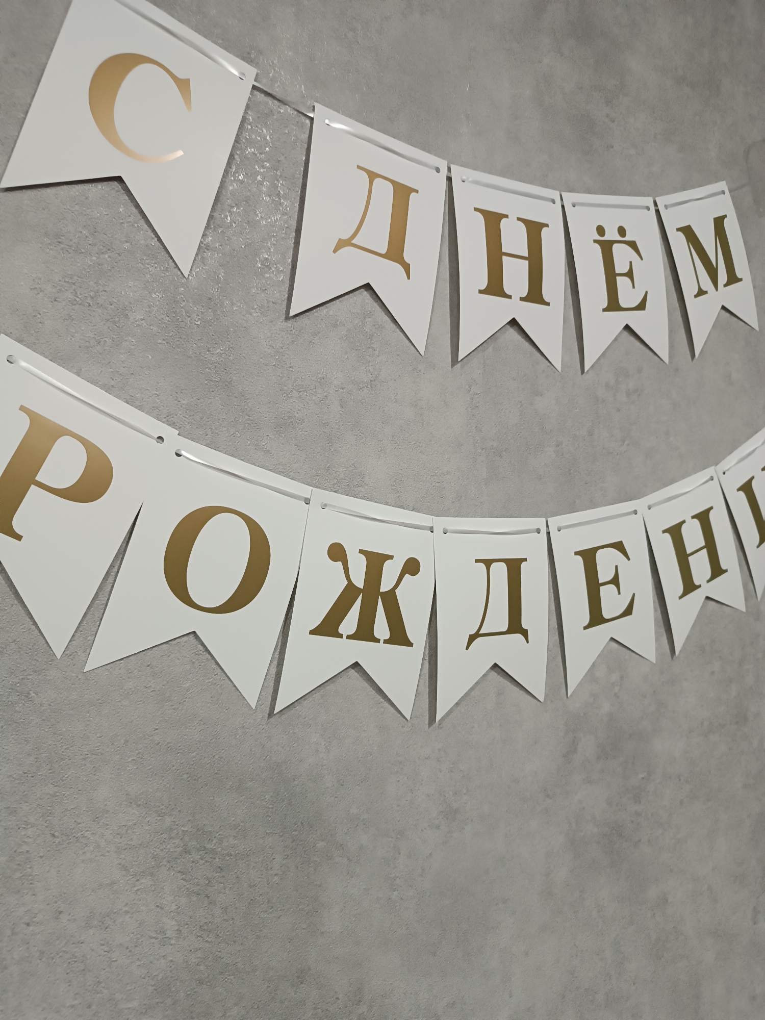 A two-line birthday banner, displaying the Russian phrase "С днём рождения" (Happy Birthday) in gold Cyrillic letters on white, flag-shaped pennants, is affixed to a textured, light gray wall. The upper line consists of five pennants ("С днём"), and the lower line contains eight pennants ("рождения"). Each pennant is strung horizontally on a thin, clear line. The wall exhibits an uneven, possibly stucco or concrete finish. No individuals, actions, or interactions are visible in the scene. The location is an indoor space in Unknown, Unknown.