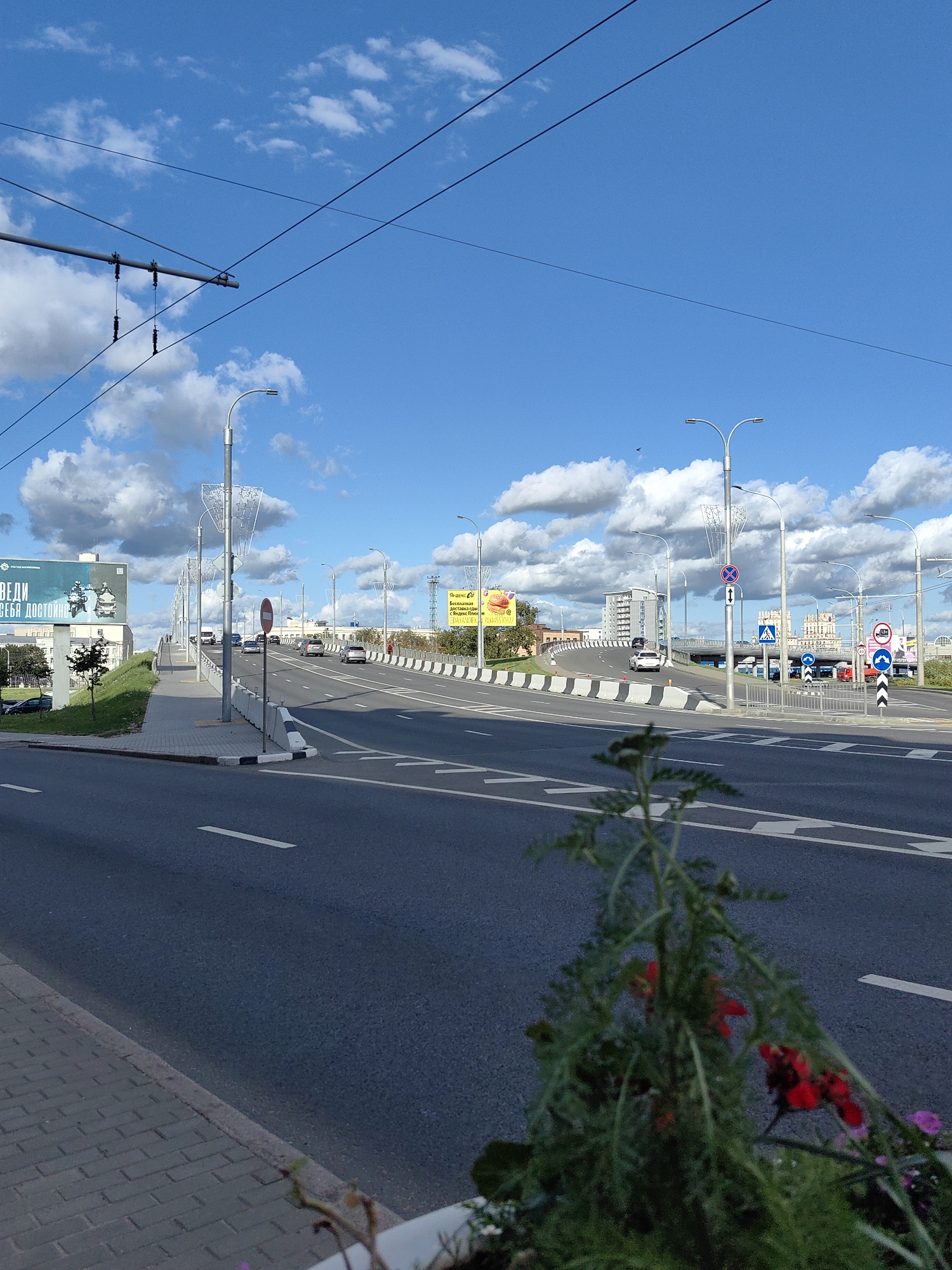The image depicts a sunny day with a clear blue sky and scattered clouds over a city street in Minsk, Belarus. The foreground features a paved sidewalk with a planter containing a flowering plant. Beyond the sidewalk, a multi-lane road curves and ascends over a bridge or elevated section. Several cars are visible on the road, moving in various directions. Streetlights line the road, and overhead power lines are prominent. Billboards and buildings are visible in the background. A red and white striped barrier is present along the side of the road, and various traffic signs, including a pedestrian crossing sign and a prohibition sign, are visible. The scene appears to be a busy urban thoroughfare.
