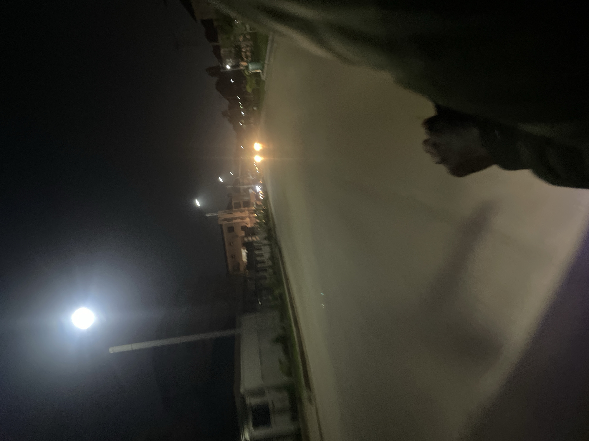 The image depicts a nighttime scene on a paved road, likely in the Guzape district of Abuja, Nigeria. The lighting suggests an urban or suburban environment, with streetlights illuminating the road. In the foreground, a portion of a person's shoulder and arm is visible, indicating the photo was taken from the perspective of someone on a moving vehicle or stationary object.
The road appears to be a two-lane street with a divider or shoulder. Buildings, some with visible lights, are present on either side of the road. On the left side, there is a building with a light-colored facade and a fence. On the right side, several structures, possibly houses or commercial buildings, are visible, some with architectural details suggesting modern construction. Trees and vegetation are also present along the roadside.
The sky is dark, consistent with nighttime. A bright streetlight is prominent in the upper left quadrant of the image, its light casting a glow on the surroundings. In the distance, headlights from an oncoming vehicle are visible, their beams cutting through the darkness. The overall atmosphere is that of a quiet, possibly residential or mixed-use area at night. The image is somewhat blurry, particularly in the foreground, suggesting movement during the capture.