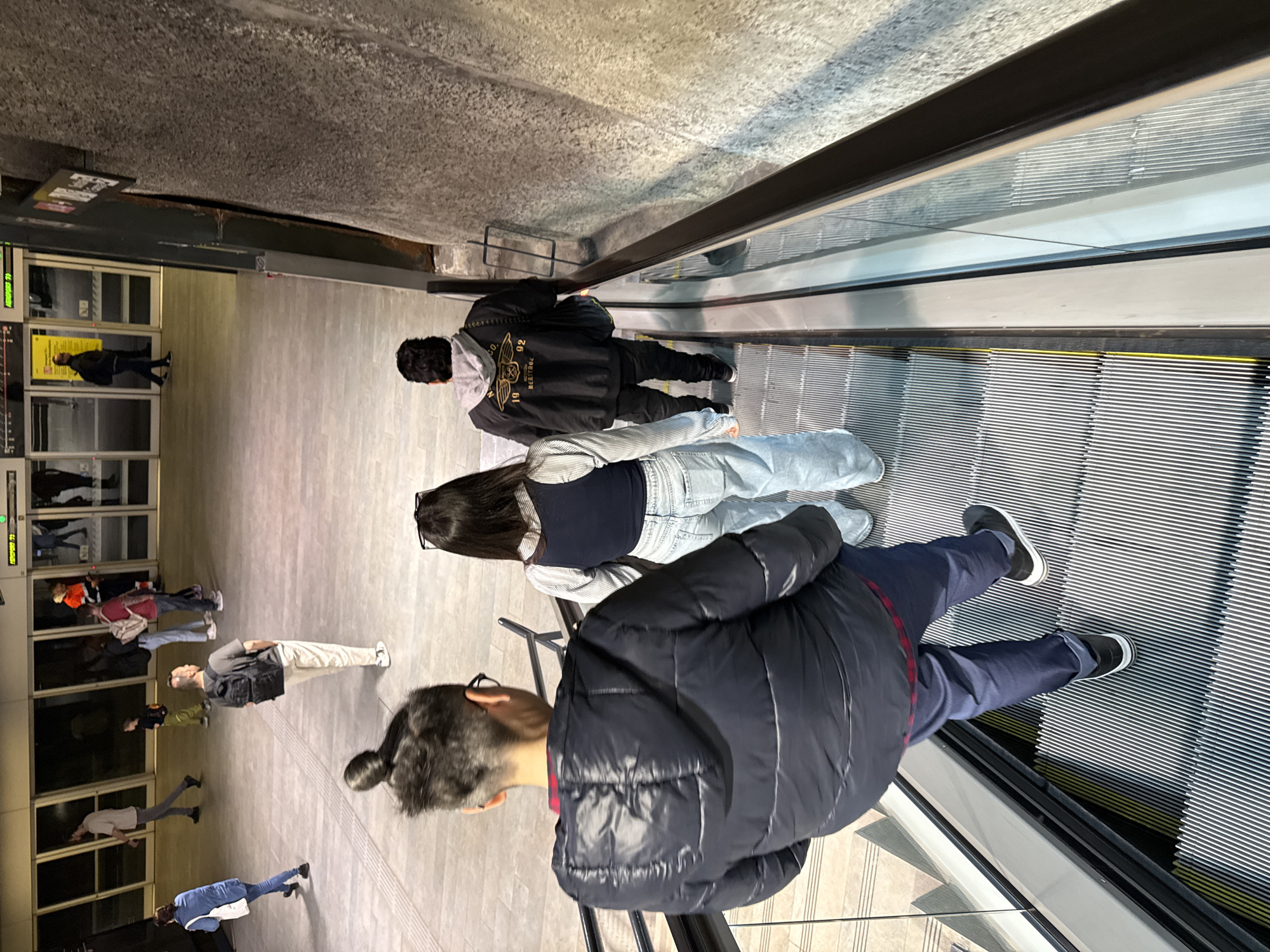 The image shows three people riding an escalator in l'Hospitalet de Llobregat, Spain. The escalator is descending. One person is wearing a dark blue puffy jacket and blue pants. Another is wearing a dark shirt and light blue jeans. A third person is wearing a black jacket with a grey hoodie and dark pants. In the background are other people walking on a light grey floor toward glass doors.