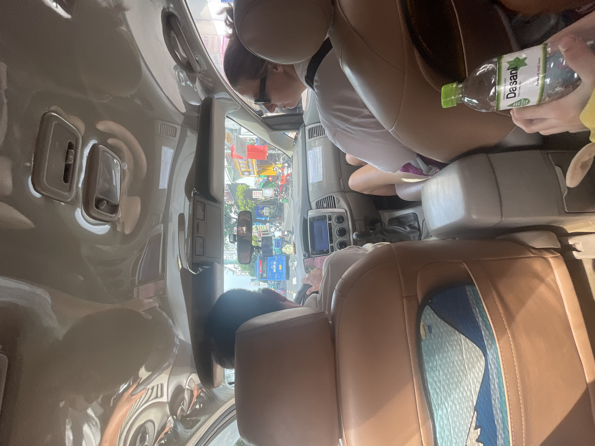 The image depicts the interior of a car, likely a minivan, viewed from the back seats looking forward. Two people are visible. In the front passenger seat, a person is wearing glasses and a white shirt, looking down. To their right, another person's hand is holding a green-capped bottle of Dasani water. The driver is visible in the driver's seat, with their hands on the steering wheel, looking forward at the road.
The car is in motion, as indicated by the blurred background of a busy street scene. Traffic, including motorcycles and cars, is visible through the windshield. A blue bus with Vietnamese license plates ("50D-207.28") is in front of the car. The street appears to be in a city environment, with buildings and signage, suggesting an urban setting. The sunlight suggests it is daytime, and the overall atmosphere appears to be one of casual travel. The interior of the car features beige leather seats and a central console. A colorful patterned object is visible in the seat pocket of the rear seat.
Visible text includes "Dasani" on the water bottle and Vietnamese characters on the street signs and the bus. The scene is set in Thủ Đức, Vietnam.