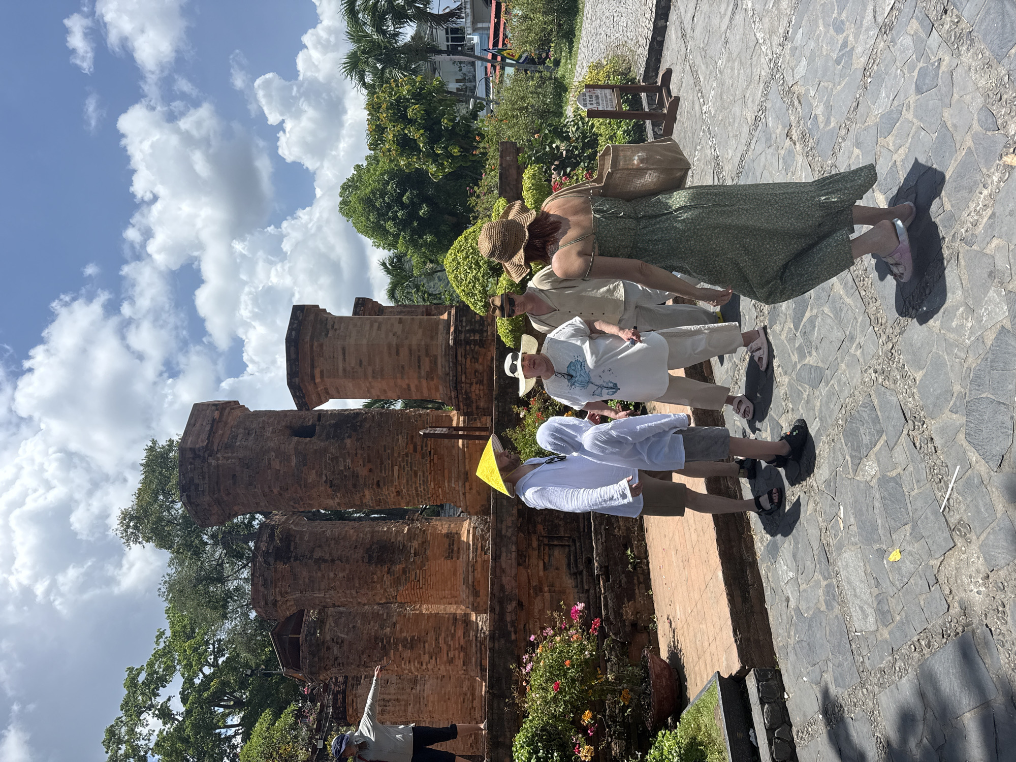 The scene is located in the city of Bac Nha Trang Ward, Vietnam. Four people are on a stone walkway that leads to brick structures. The first person is wearing a yellow hat, white shirt, and brown shorts. The second person is wearing a white hat, white t-shirt with blue designs, and white pants. The third person is wearing a tan shirt, brown pants, and sunglasses. The fourth person is wearing a green dress and a straw hat. In the background are brick structures, trees, and a partially cloudy sky. A man is standing in front of a flower bed with his arm raised.