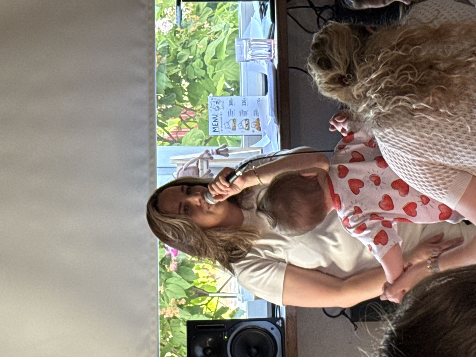 An indoor scene in Balashikha, Russia, captures a woman speaking into a microphone while a child interacts with her. The primary subject is an adult female, positioned centrally, with light brown, wavy hair. She wears a plain, light-colored short-sleeved t-shirt and a gold-toned bracelet on her left wrist. Her right hand holds a black microphone, positioned near her mouth as if speaking or singing.
A young child, observed from the back, is positioned in front of the woman, slightly to her left. The child wears a white t-shirt adorned with red heart patterns. The child's right arm is raised, hand extended towards the microphone or the woman's hand.
The background features a large window, partially obscured by a light-colored roller blind pulled down from the top. Through the uncovered portion of the window, lush green foliage and scattered light pink flowers are visible, suggesting an outdoor garden or natural setting.
Beneath the window, a wooden surface functions as a shelf or counter. On the left side of this surface, a black audio monitor speaker is visible. To the right of the speaker, a white menu card stands upright, displaying simple illustrations of cat-themed sandwiches and prices listed as "300p" and "330p", indicating Russian Rubles. Adjacent to the menu, a clear glass filled with water and several loose white papers are present. A light-colored, multi-limbed plush object hangs near the window frame on the left side of the menu.
In the foreground, partially visible individuals observe the scene. On the far left, the back of an adult's head with dark hair and a forearms with a multi-band bracelet are seen. On the far right, the back of an adult woman's head and upper torso are visible. She has curly, light-colored hair, seemingly tied up, and wears a cream-colored, textured knit top. Black cables are partially visible behind this individual and the speaker. The overall lighting is bright, suggesting natural light from the window.