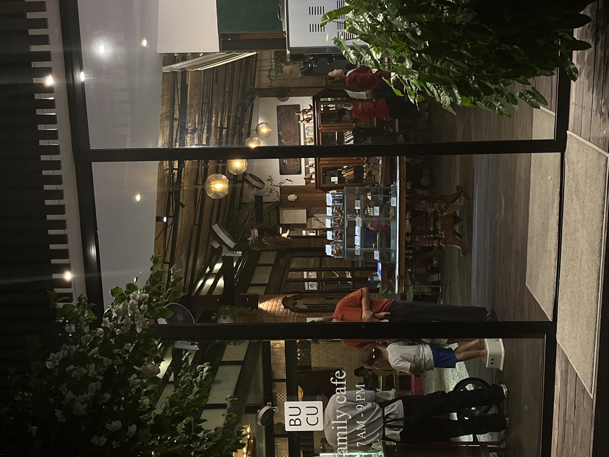The image captures the exterior view of a cafe named "BU CU Family Cafe" in North Kuta, Indonesia, at night. The cafe's interior is visible through large glass doors, revealing a warm, dimly lit space with wooden floors and a rustic ceiling structure adorned with industrial-style pendant lights. Several people are seen inside, including what appears to be staff members in uniform and customers. A man in a grey uniform with a black apron is standing near the entrance, facing away from the camera. A young child in blue shorts and a white t-shirt is standing on a white stool outside the cafe, looking into the interior. Another person in an orange shirt is standing further inside. Display cases filled with what seem to be food items or merchandise are visible. Outside the cafe, lush green plants frame the right side of the image. The overall scene suggests a welcoming and relaxed atmosphere of a family-friendly establishment operating after dark.