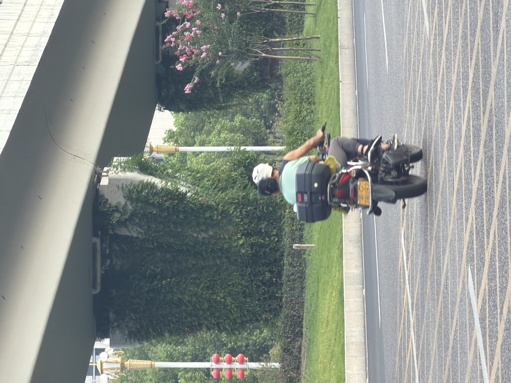 The image shows two people riding on a motorcycle on a street in Jiangjin, China. The motorcycle is red and black and has a black storage box on the back. The person in the front is wearing a white helmet and a light green t-shirt. The person in the back is wearing a dark shirt and grey pants. They are both facing forward. The motorcycle is driving on a paved road with yellow and white markings. In the background, there are trees and a large concrete overpass. There are also some red lanterns on a pole to the left of the image. The scene appears to be in an urban environment.