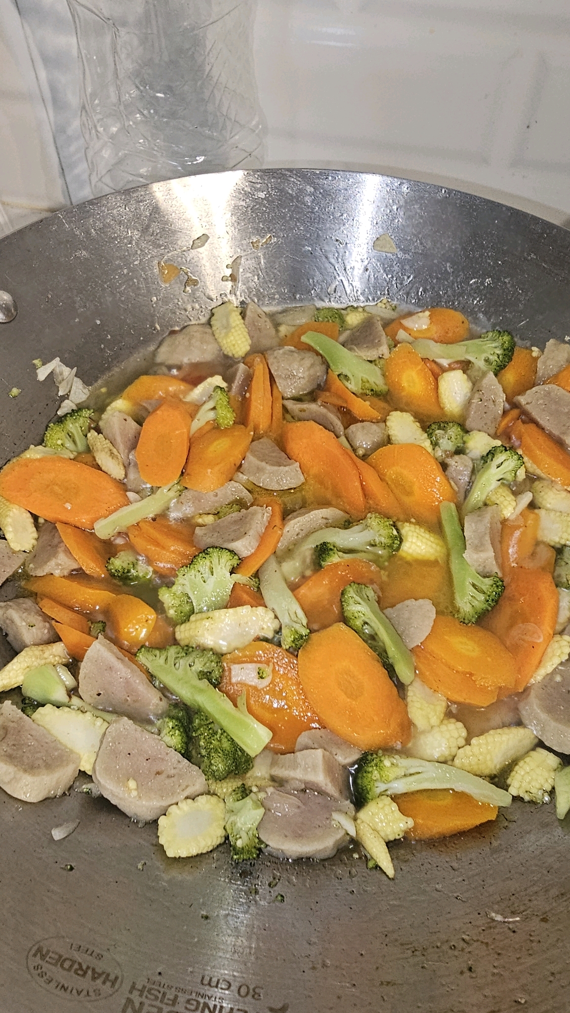 A close-up, overhead shot captures a wok filled with a vibrant stir-fry in Cibinong, Indonesia. The dish features sliced carrots, florets of broccoli, baby corn, and chunks of meat, all simmering in a light broth. The wok itself is made of stainless steel and bears visible signs of use, with some discoloration and remnants of food stuck to its surface. The background is out of focus, showing a plastic bottle and a white wall. The overall impression is of a home-cooked meal prepared in a typical Indonesian kitchen.